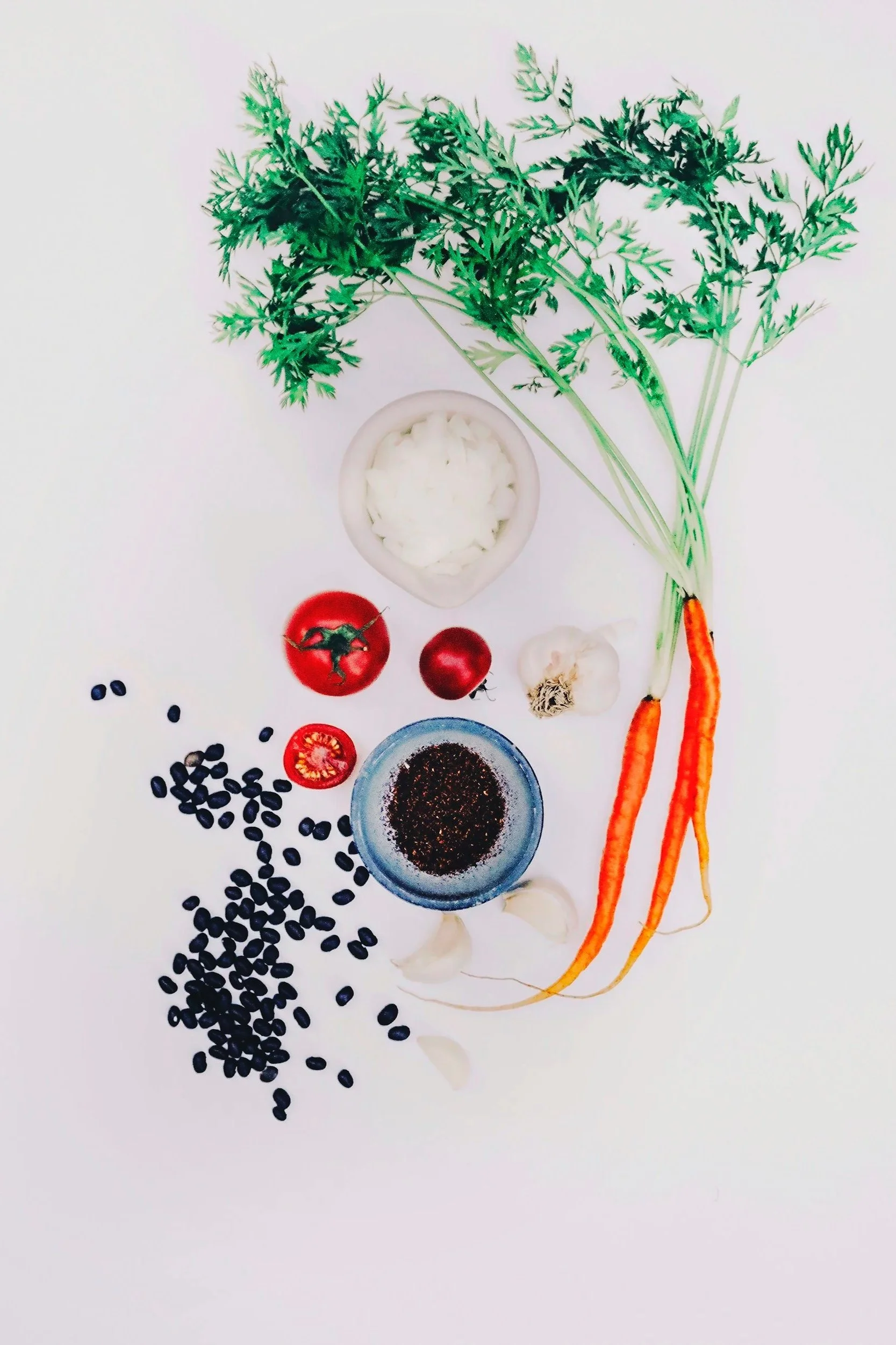 Fresh parsley, cherry tomatoes, garlic, black beans, onion powder, black pepper, and carrots arranged on a white surface.