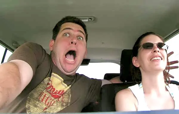 Man and woman in car, taking a selfie. Man making a surprised or funny face, woman smiling.