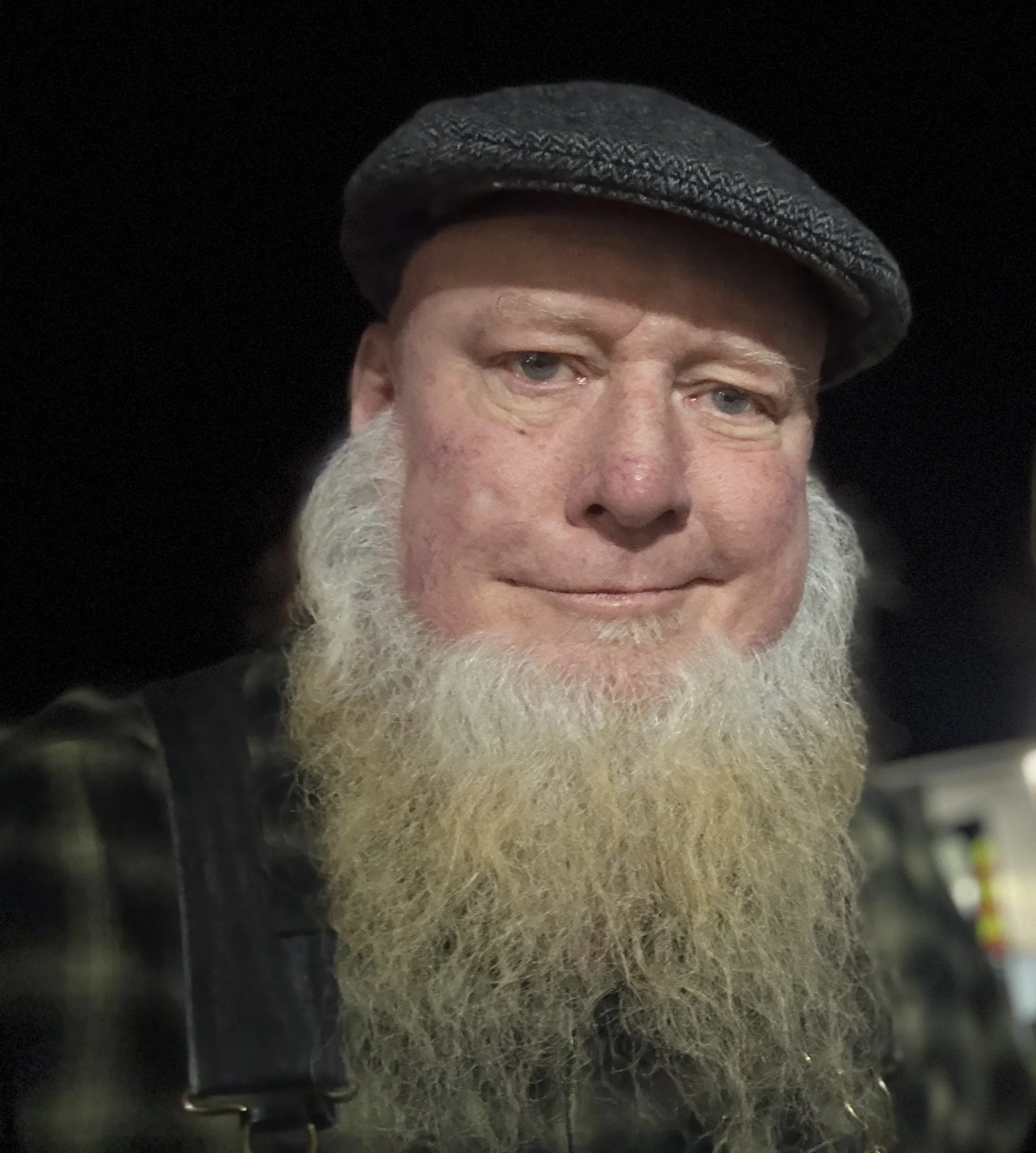 A close-up of a man with gray hair and beard wearing a black flat cap, with a dark background.