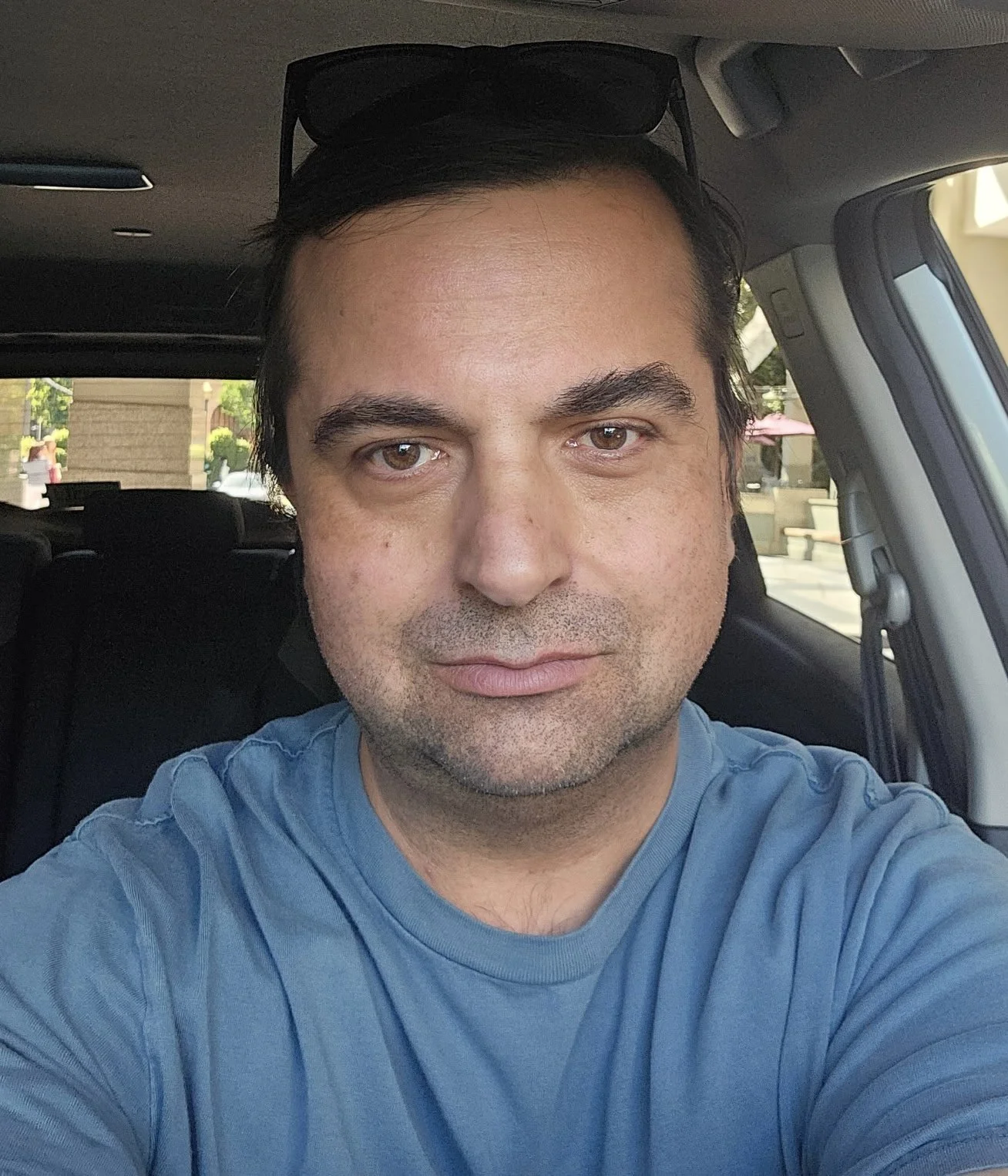 A close-up selfie of a man with dark hair, light skin, and brown eyes, wearing a blue t-shirt and sunglasses resting on his head, sitting inside a vehicle with a window showing an outdoor scene.