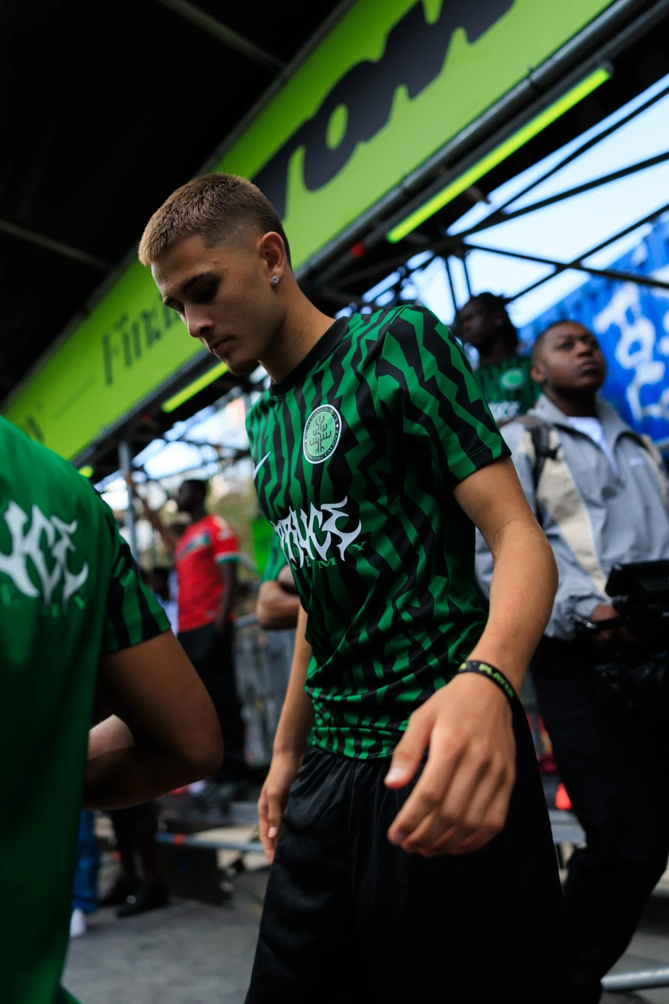 Close-up of a young male athlete with short hair, wearing a black and green sports jersey, standing in an outdoor setting with other people in the background.