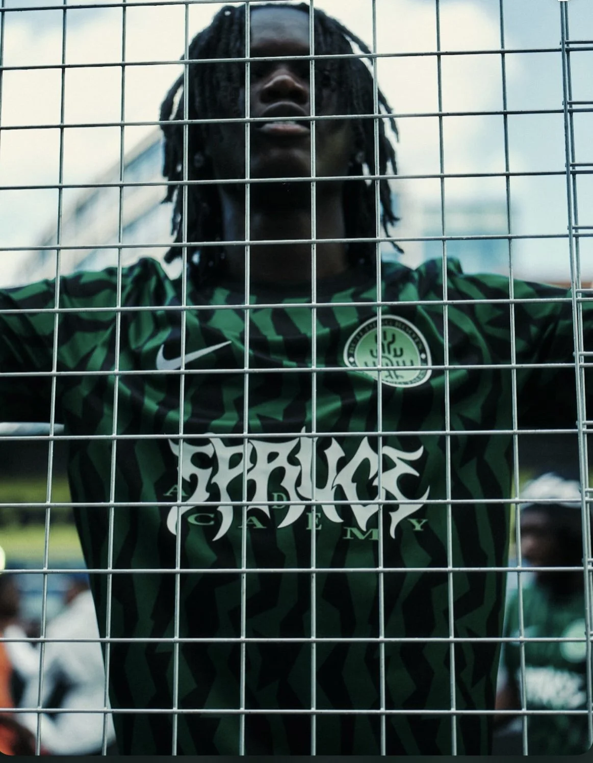 A person with dreadlocks wearing a green and black sports jersey behind a metal fence.
