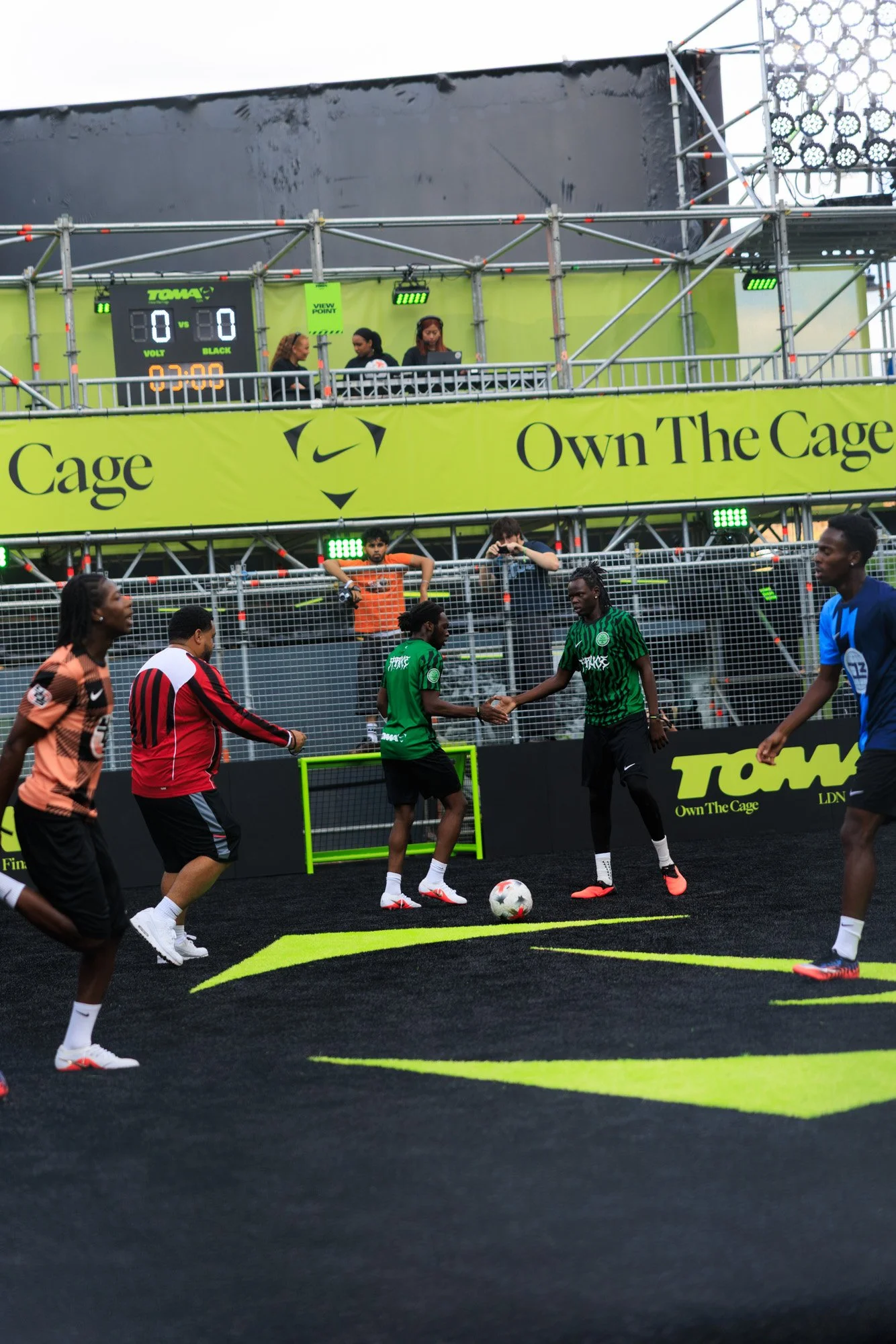 Soccer players warming up or practicing on a black field with neon yellow markings, with a barrier fence behind them and a yellow banner that says 'Cage' and 'Own The Cage.' There are spectators and officials watching, and some people on the elevated
