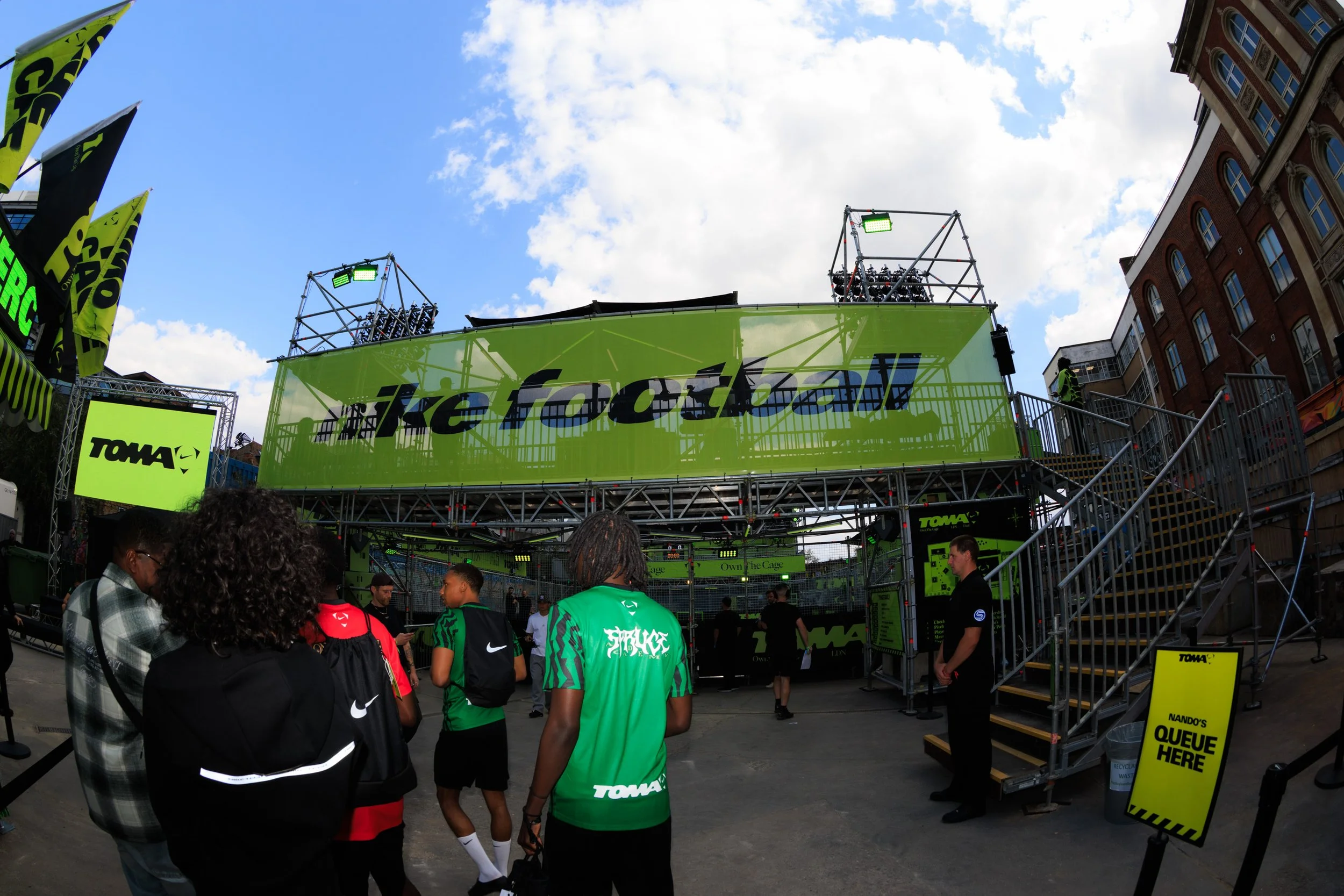 People gathered at an outdoor event with a large green and black stage structure, banners, and signs. The stage displays the TOMA logo and various screens. The sky is partly cloudy, and surrounding buildings are visible.