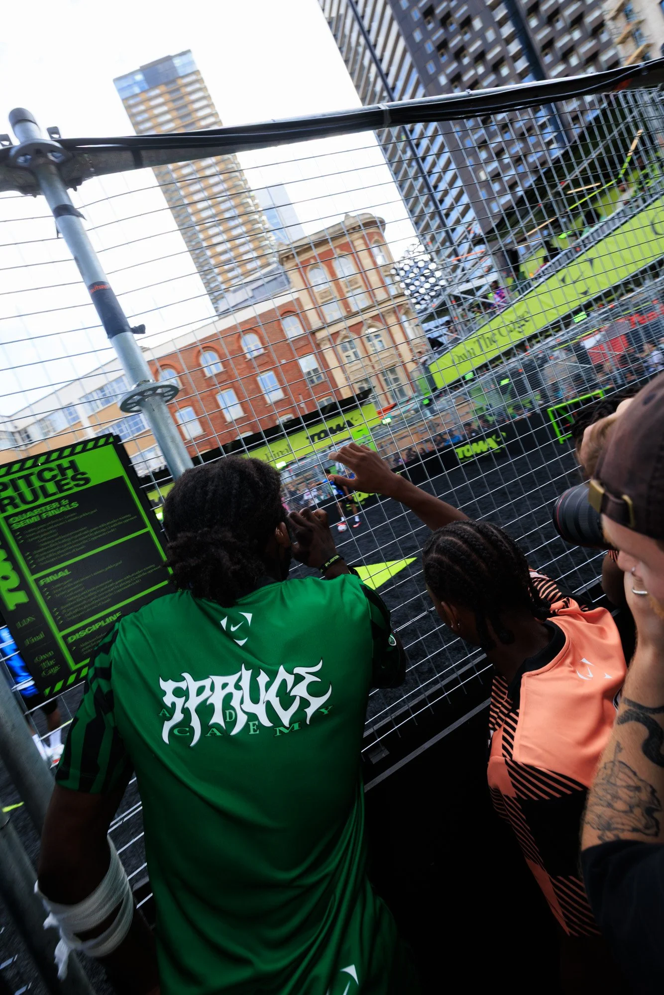 People standing behind a metal fence during a sporting event, with signage about pitch rules and semi-finals, in an urban setting with tall buildings in the background.