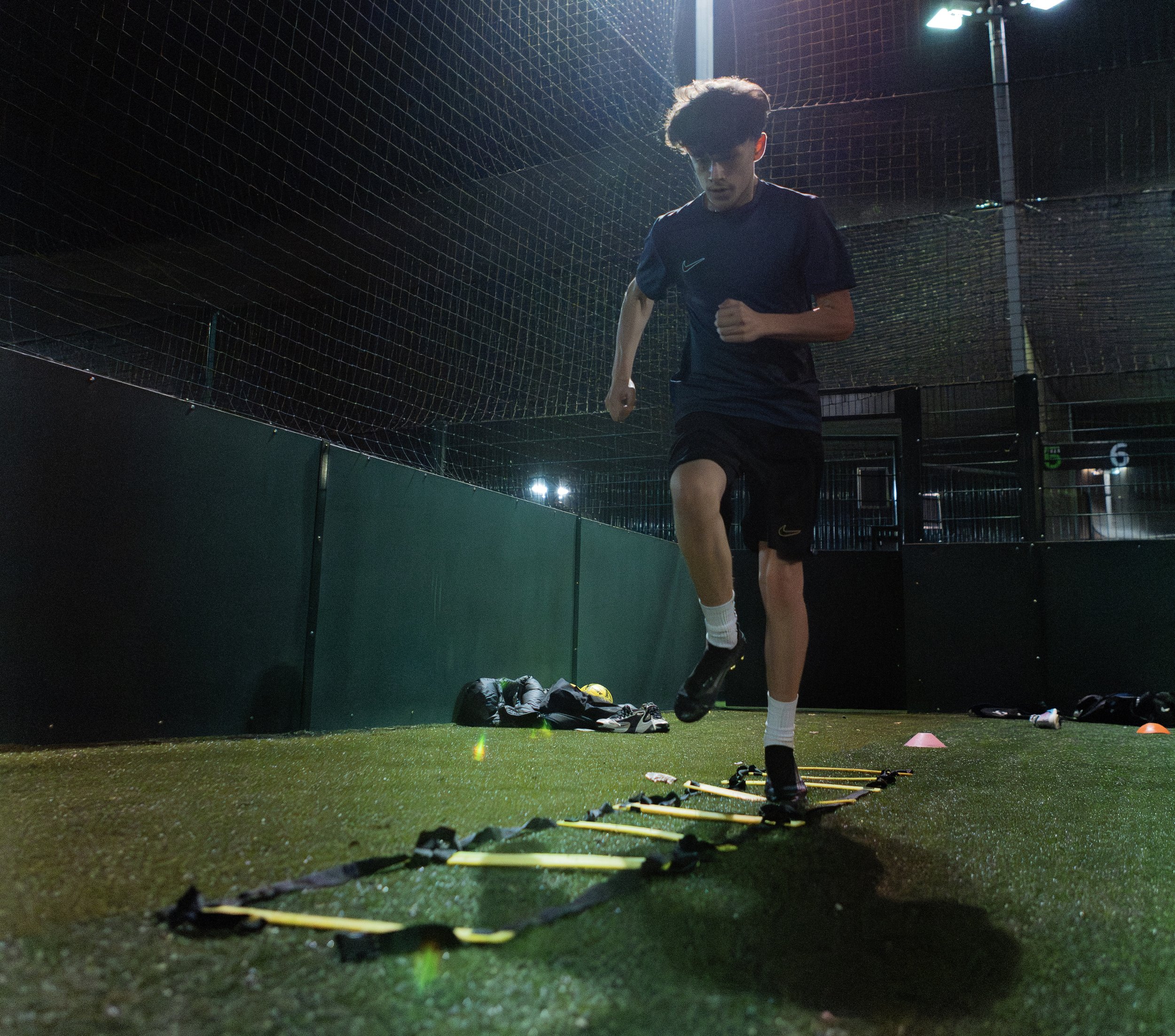 A young male athlete practicing agility drills on artificial turf at night, stepping through a yellow and black agility ladder, with sports gear and backpacks on the ground nearby.