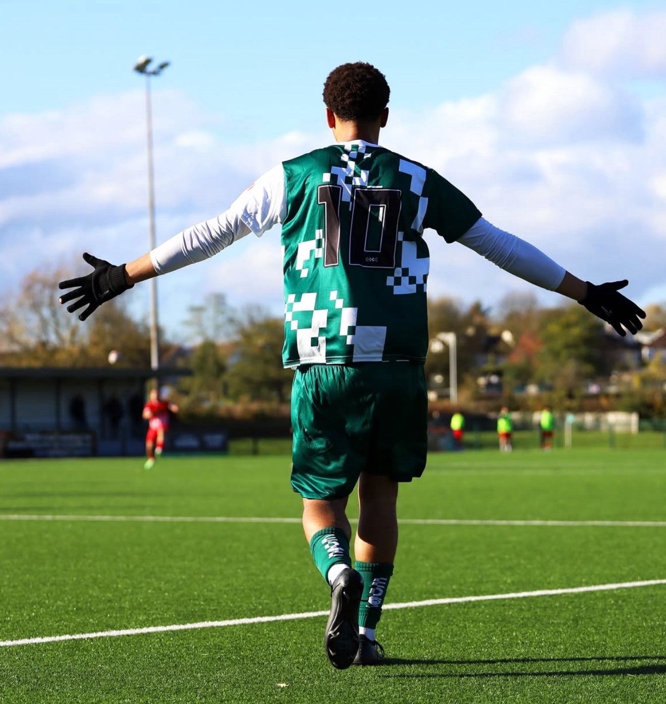 Soccer player with outstretched arms on the field, wearing a green and black jersey with the number 10, black gloves, and green shorts, with other players and a goal in the background under a partly cloudy sky.