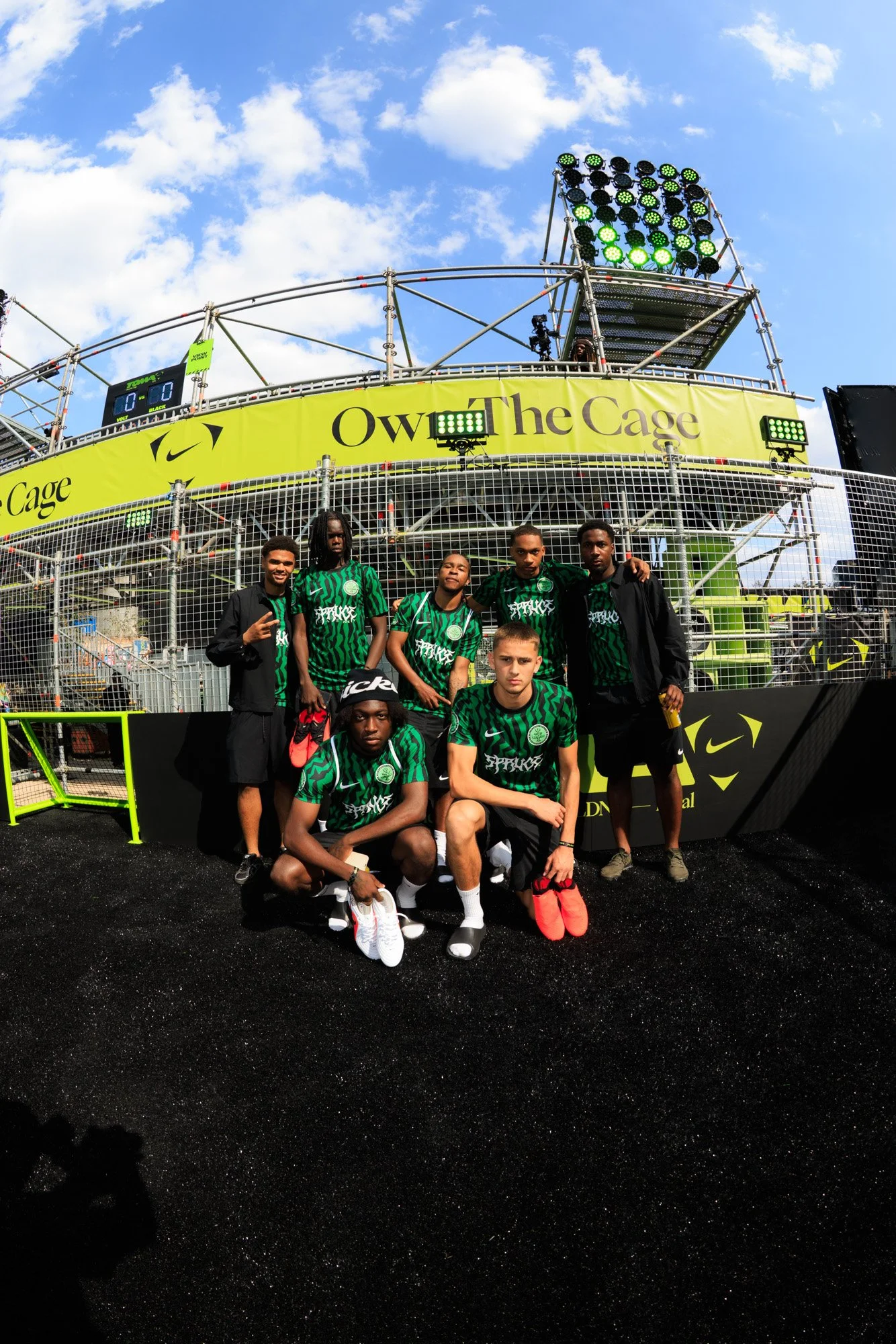A group of young men in green sports jerseys and black jackets posing for a photo in front of a yellow structure with the words "Owen The Cage." They are on a black textured surface, with a blue sky and some white clouds above.