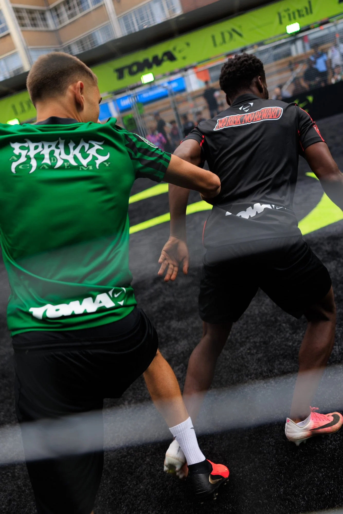 Two runners in a race, viewed from behind, running on a black track with yellow lines. The runner on the left wears a green shirt with white text and black shorts, and the runner on the right wears a black shirt with red details and black shorts. The