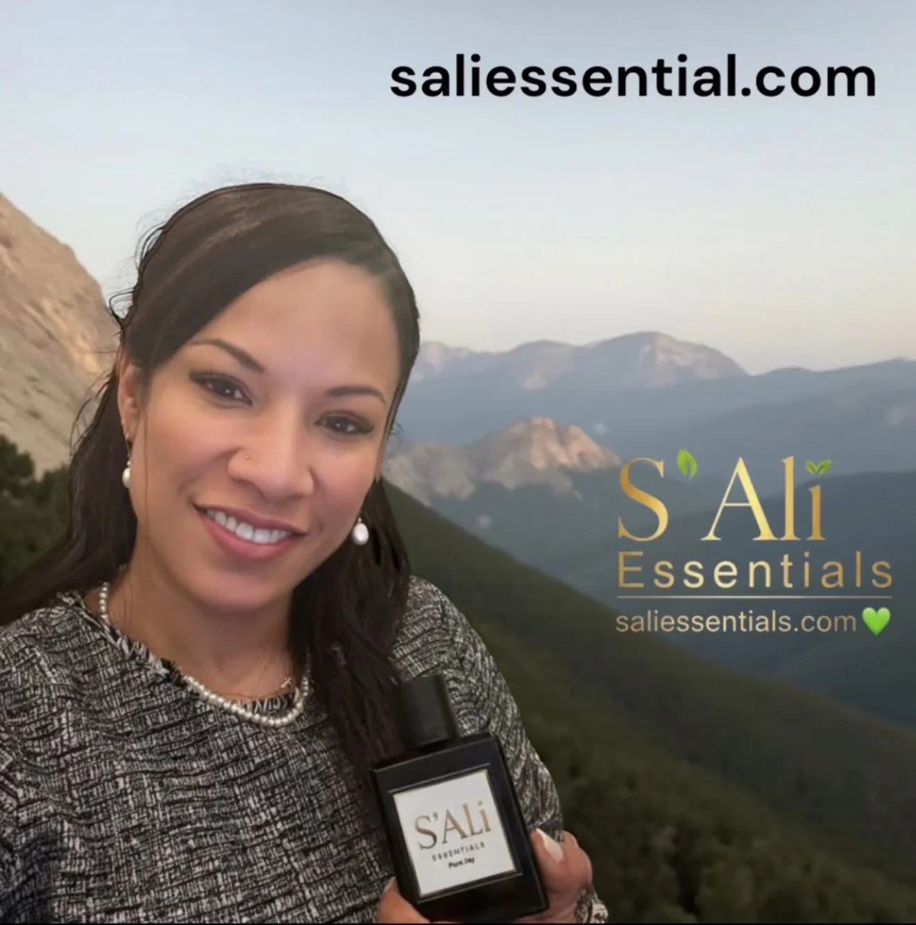 A woman smiling outdoors in front of a mountainous landscape, holding a black bottle of S'Ali Essentials perfume. Text overlays include 'saliessential.com' at the top and 'S'Ali Essentials' with a green heart at the bottom right.