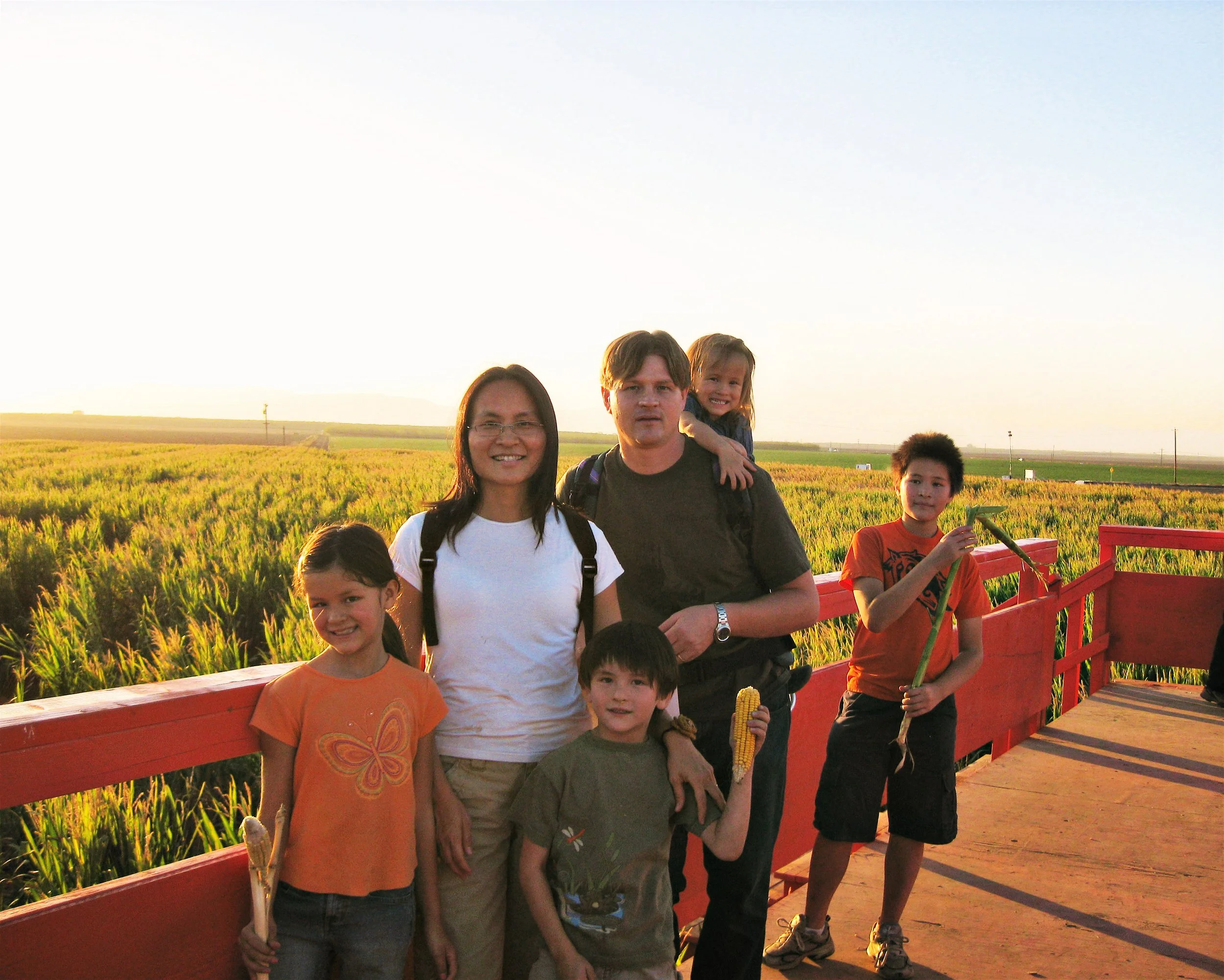 A family of six standing on a wooden platform in a field at sunset, holding corn.