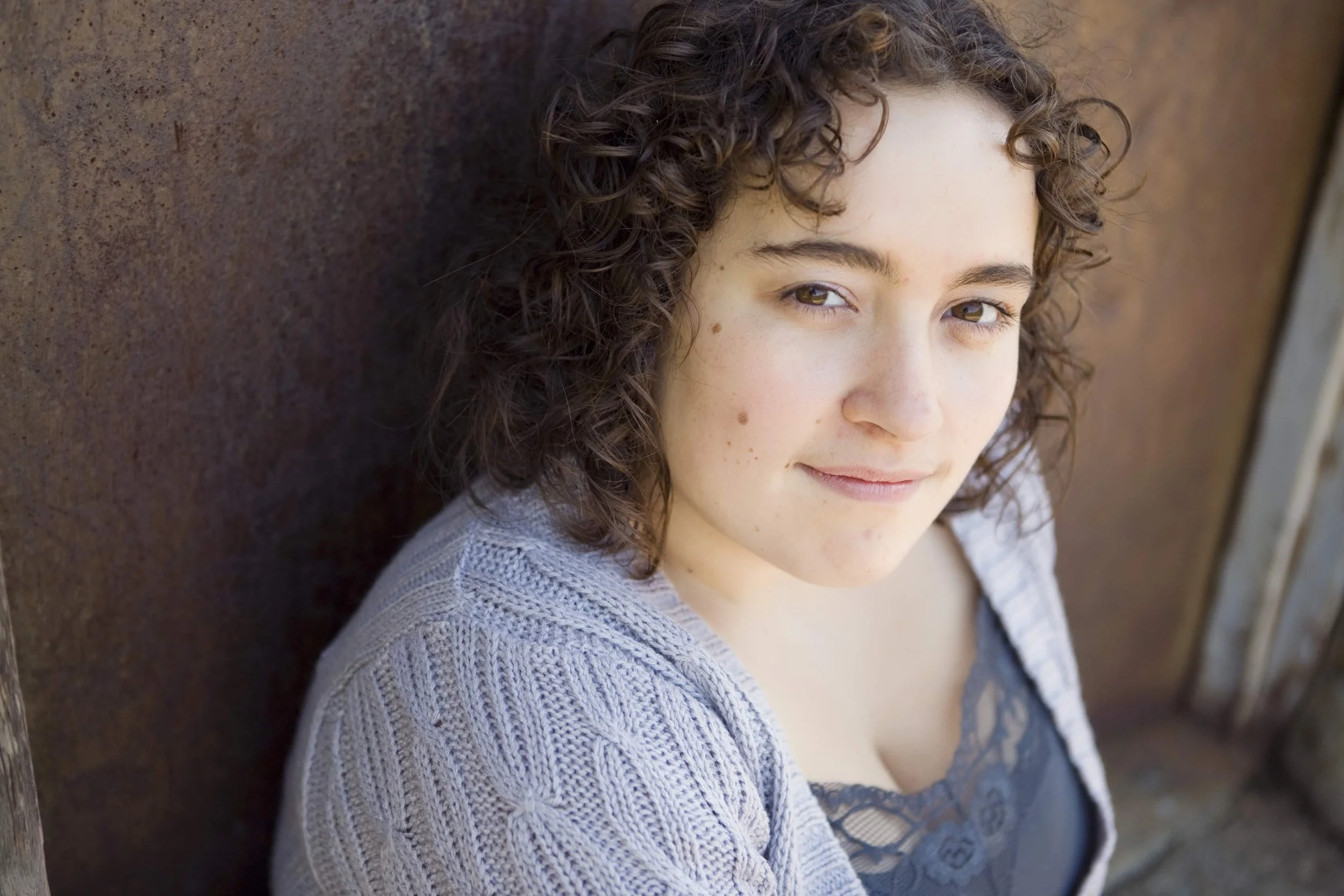A young woman with curly brown hair and light skin, wearing a gray knitted sweater and a black lace top, leaning against a rusty metal wall outdoors.