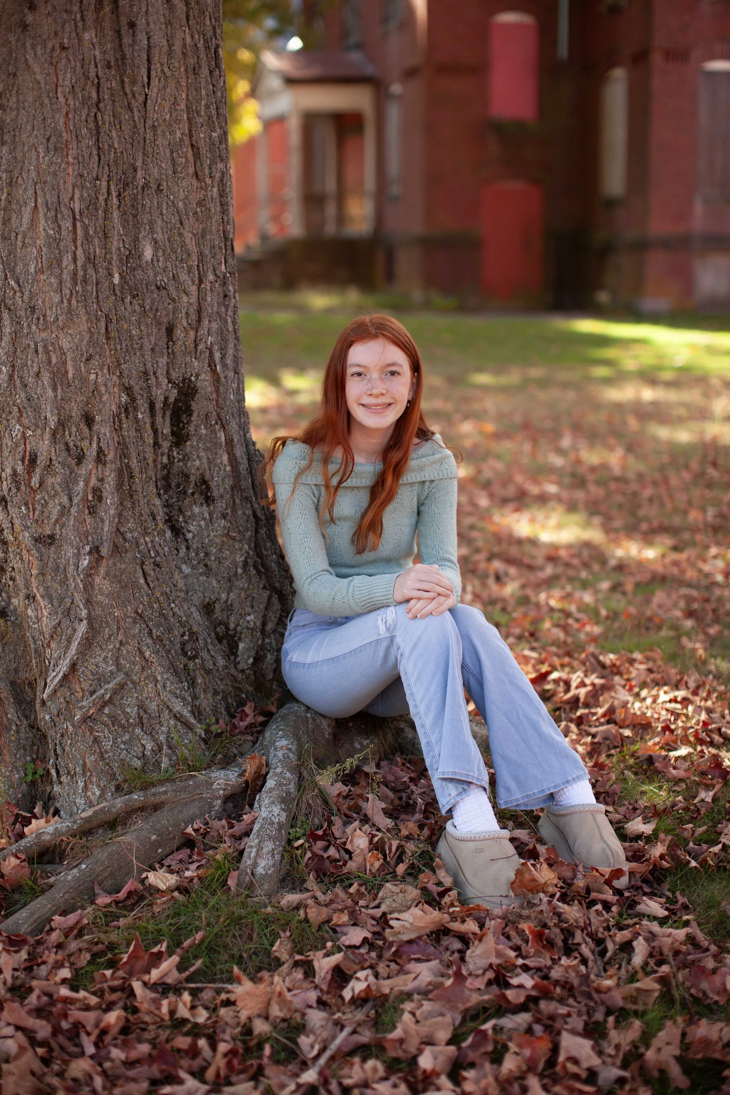 A young woman with red hair wearing a mint green sweater and light blue jeans sitting on the ground against a tree trunk, surrounded by fallen autumn leaves, with a building in the background.