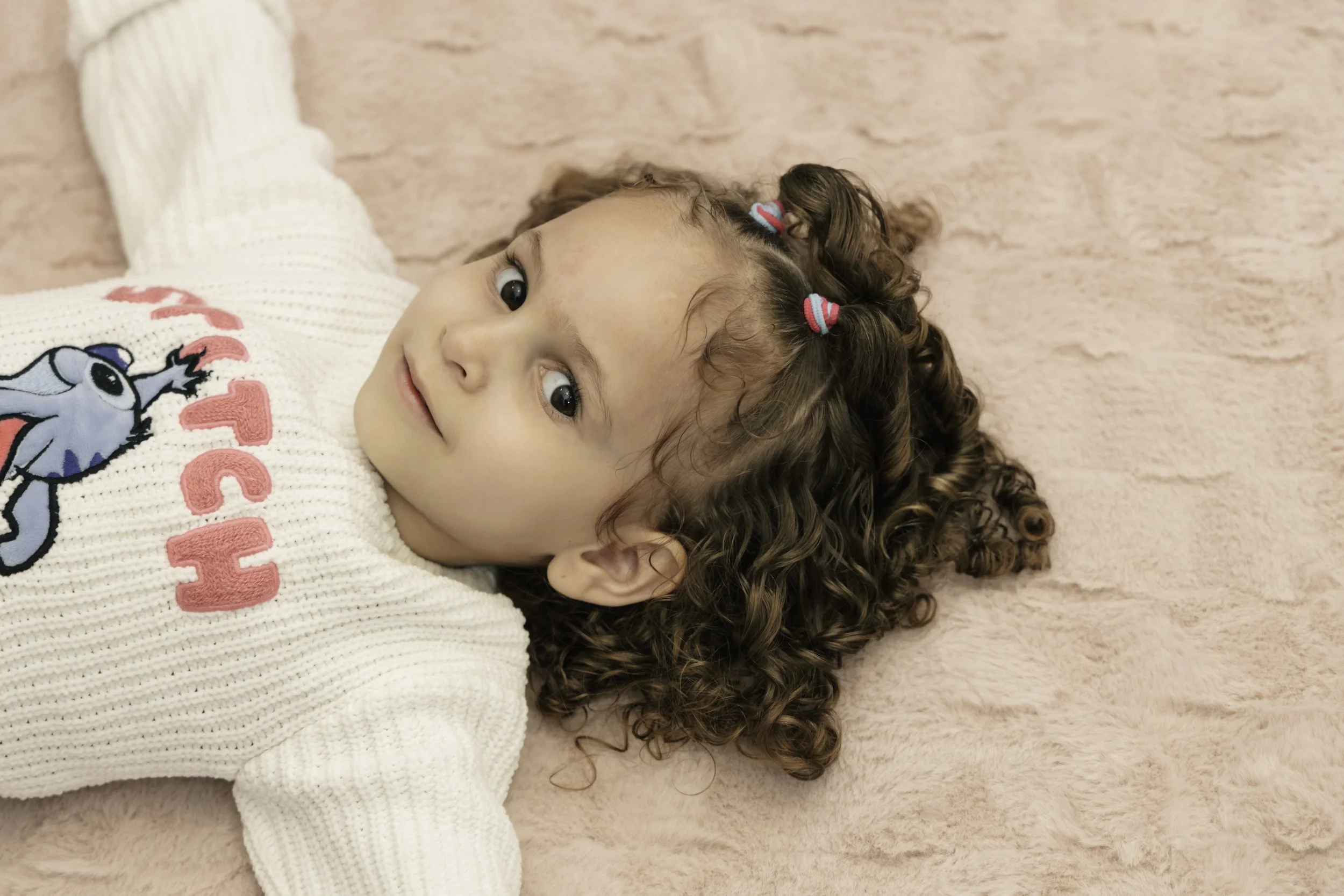 Young girl lying on a pink fuzzy blanket with curly hair held back by colorful hair ties, wearing a cream sweater with a cartoon character and pink lettering.