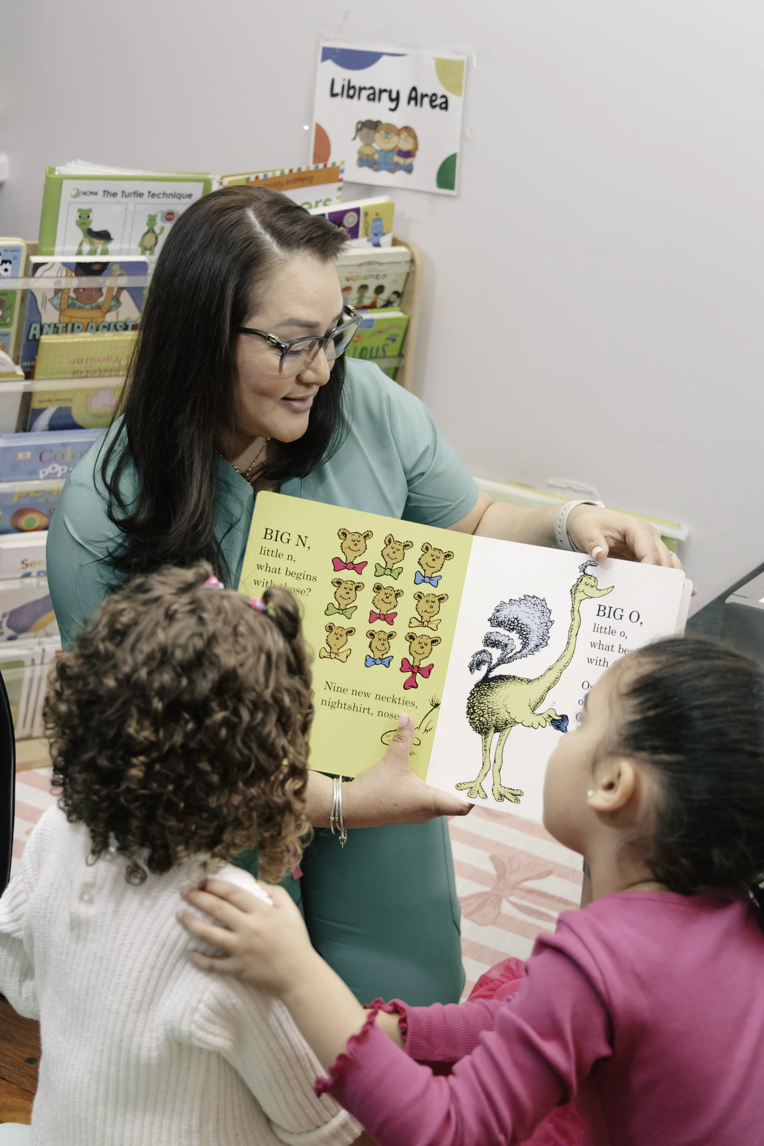 A teacher showing two young girls a picture book about animals in a library. The book shows bears with various accessories and a giraffe. There is a sign on the wall that says 'Library Area'.