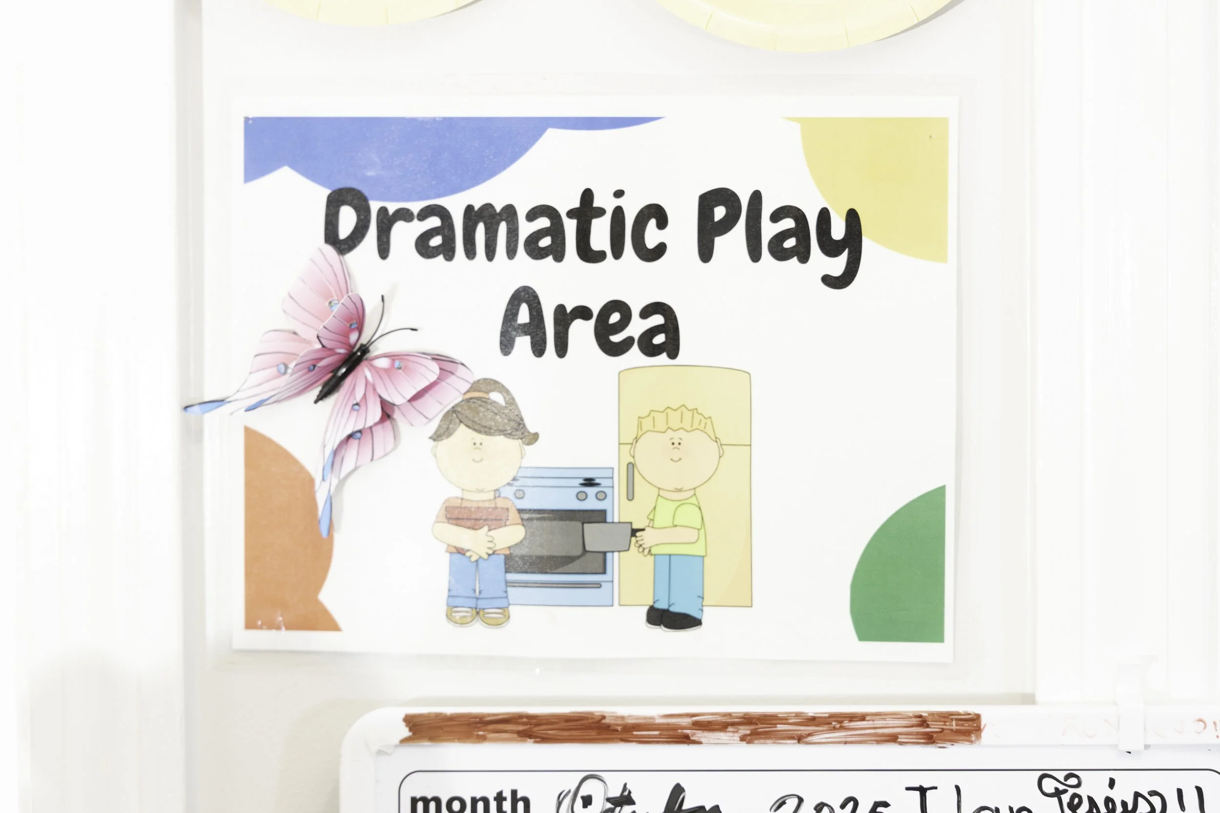 Sign reading 'Dramatic Play Area' with cartoon illustrations of two children, a girl holding a book and a boy near a stove and refrigerator, and a pink butterfly decoration.