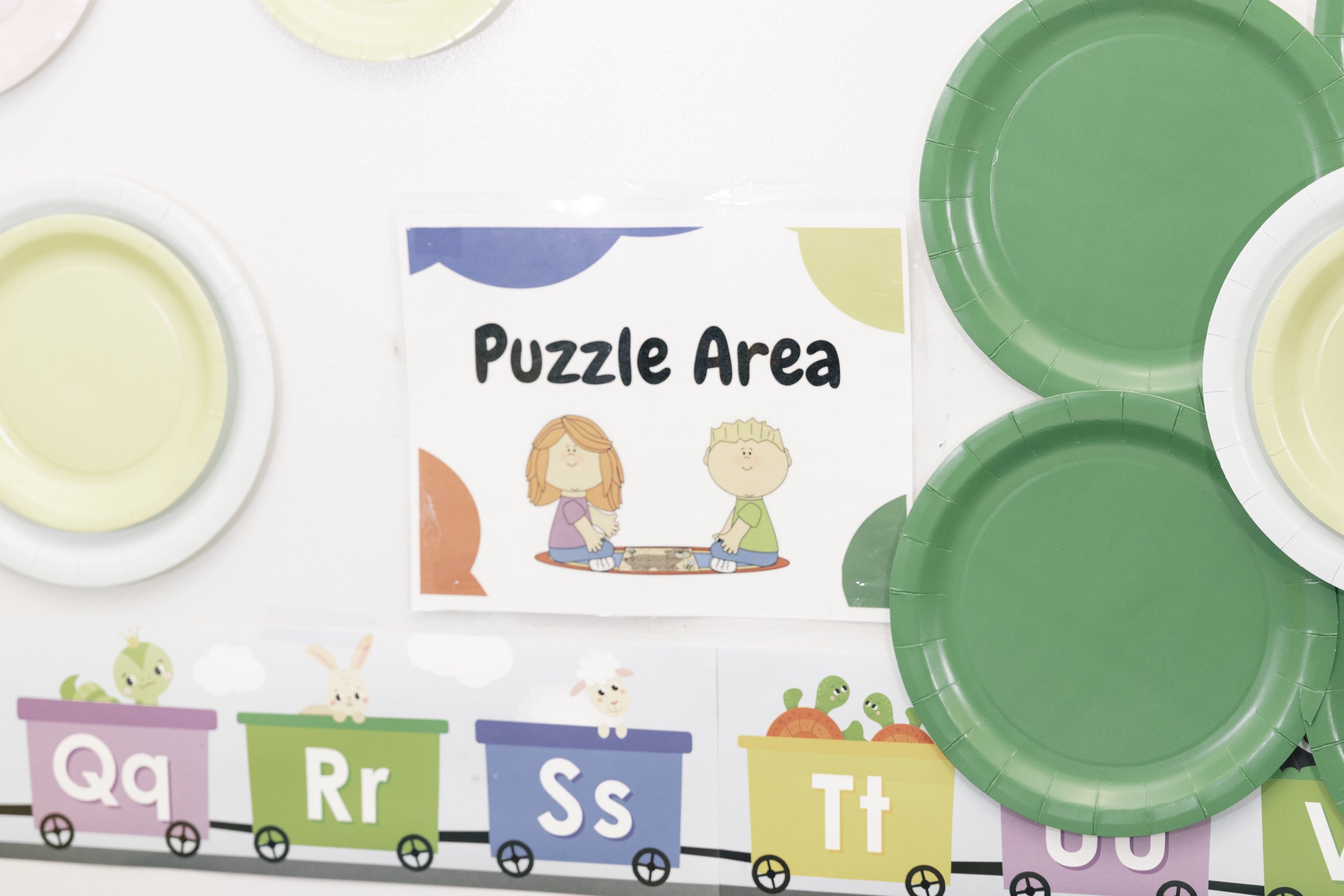 Colorful paper plates and a sign that reads 'Puzzle Area' with cartoon children playing. Alphabet-themed decoration at the bottom with multicolored carts containing animals.