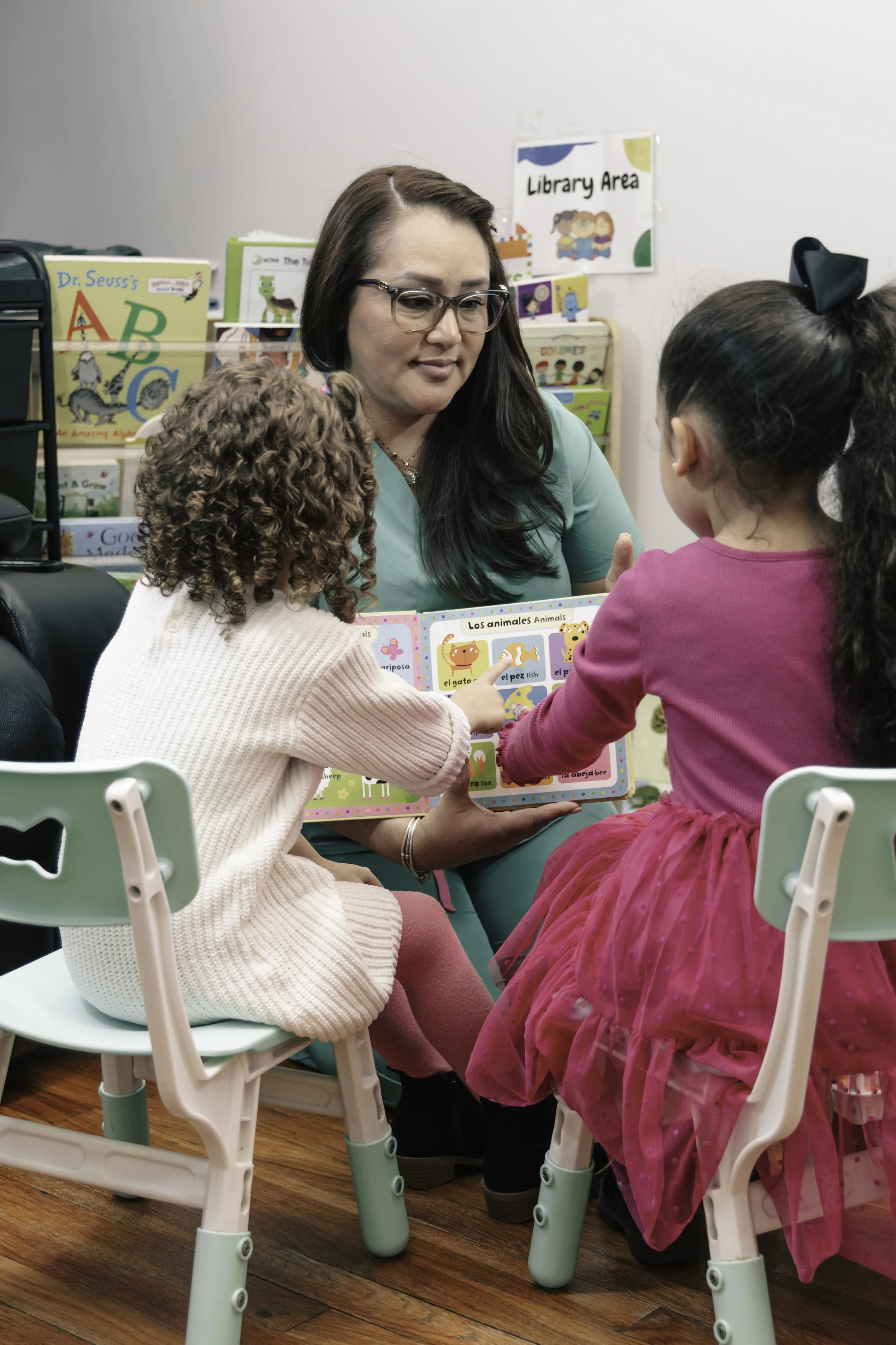 A teacher reading a children's book about animals to two young girls seated in a classroom or library. The girls are attentively looking at the book.
