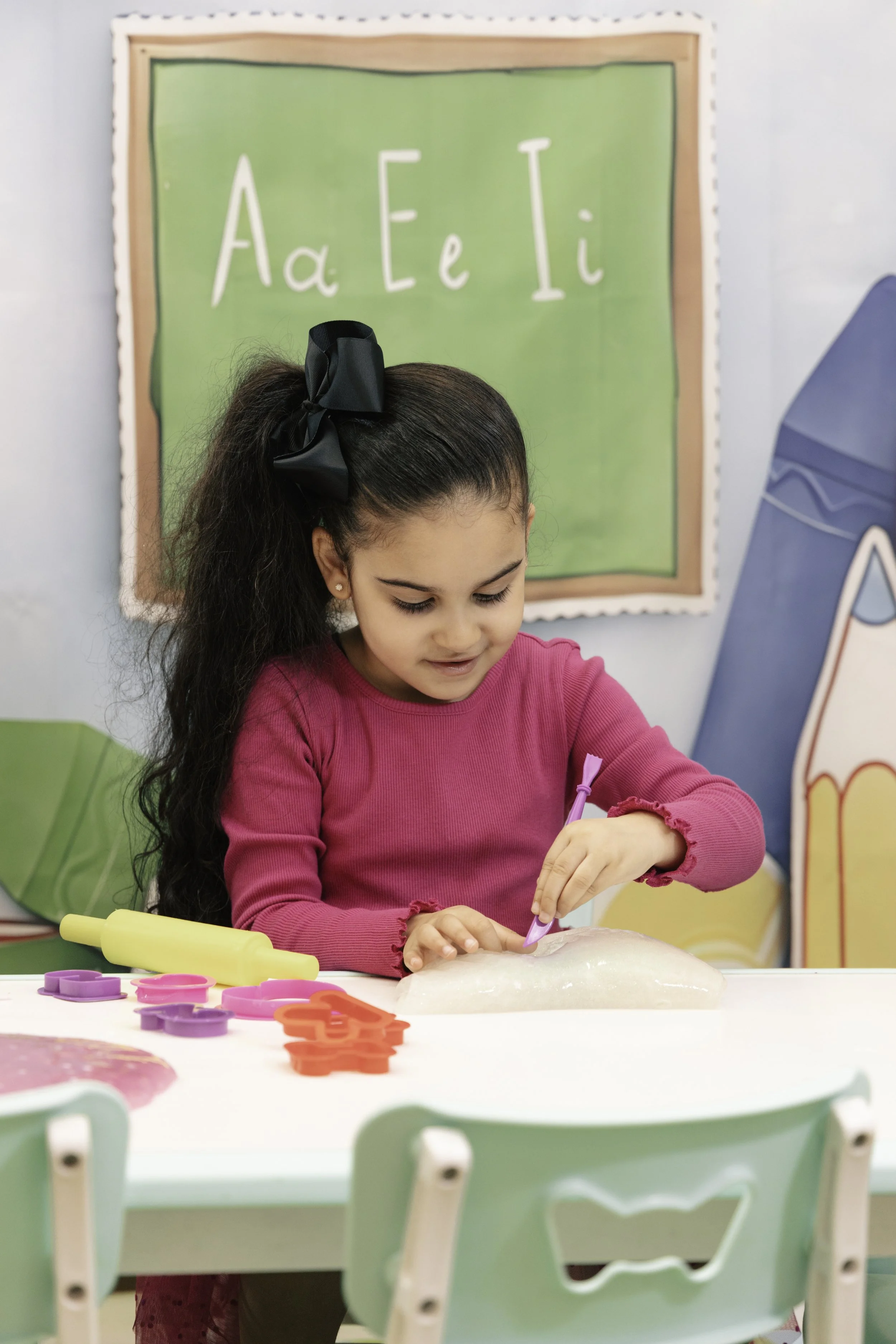 A young girl with long dark hair tied with a black bow, wearing a pink long-sleeve shirt, is sitting at a white table in a classroom, decorating a piece of dough with a purple pen. There are cookie cutters and a rolling pin on the table. In the backg
