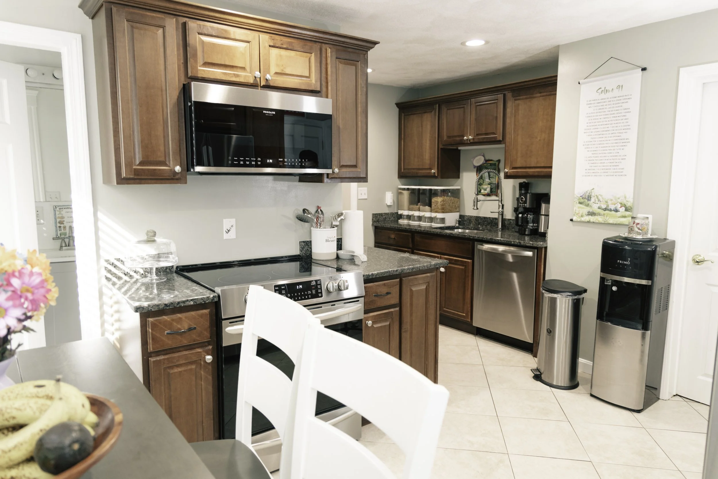 A kitchen with wooden cabinets, a microwave, stove, and stainless steel dishwasher. There are countertop containers, a coffee maker, a water cooler, and a trash can. Part of a dining table with white chairs and a bowl of bananas and avocados are visi