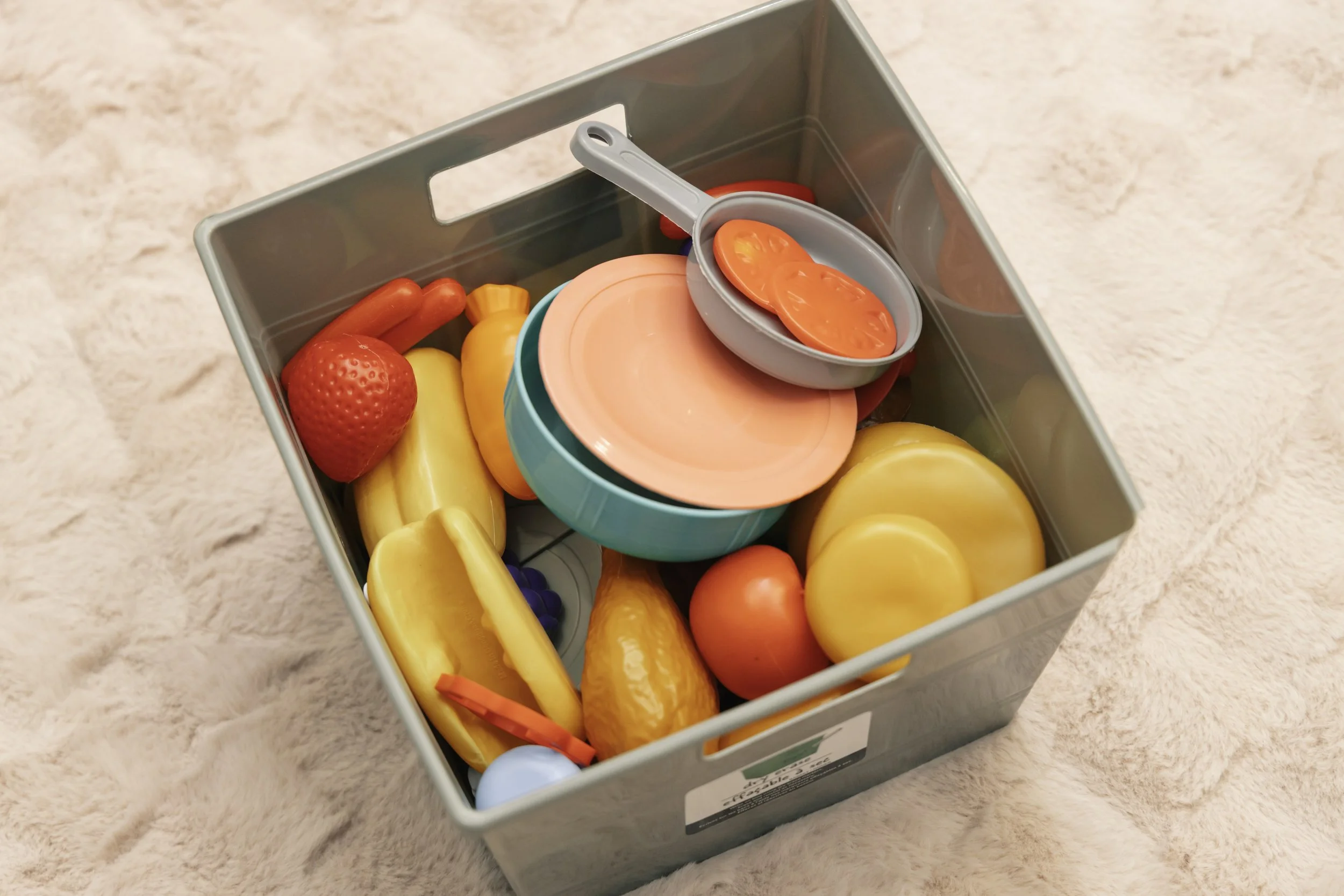 Plastic toy food set inside a gray storage bin, including toy vegetables, fruits, plates, and a pan with toy tomato slices, placed on a soft pink plush surface.