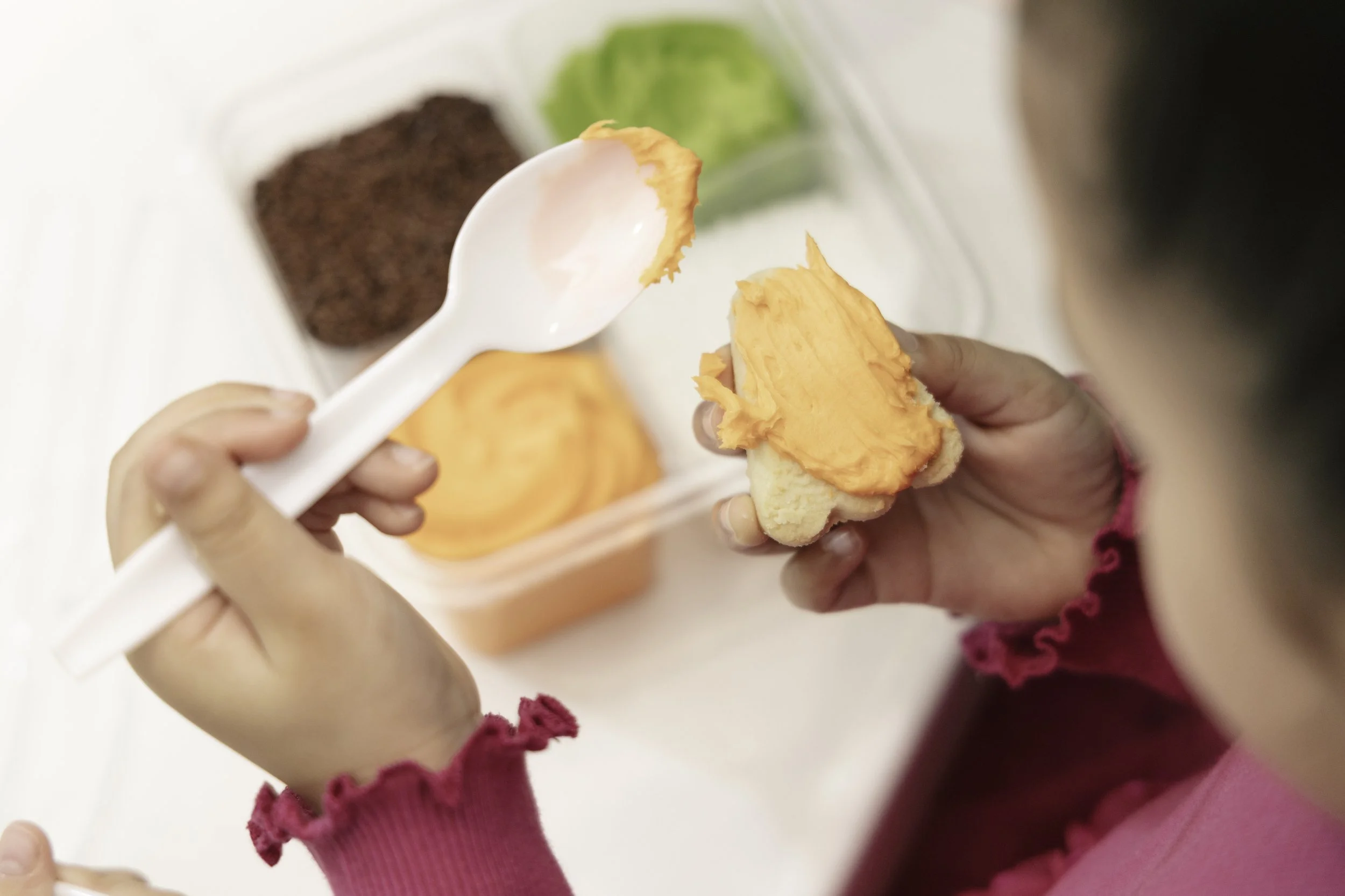 Child holding a piece of bread with orange butter, with a container of more bread and spread in the background.