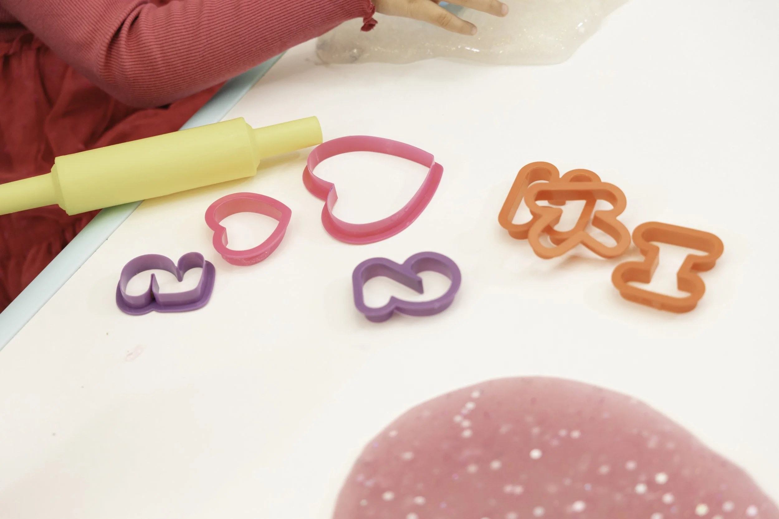 Various colorful cookie cutters in the shapes of hearts and numbers, a rolling pin, and a pink spatula on a white surface.