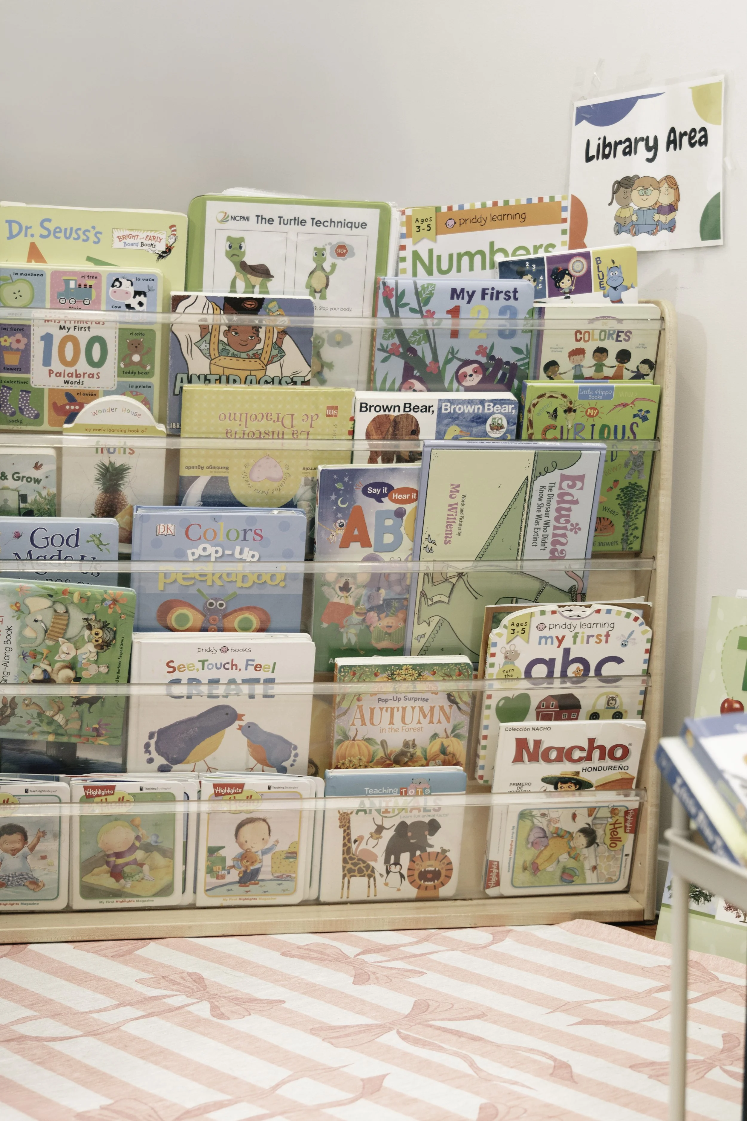 Books on a display stand in a children's library or bookstore, with a sign for the library area.