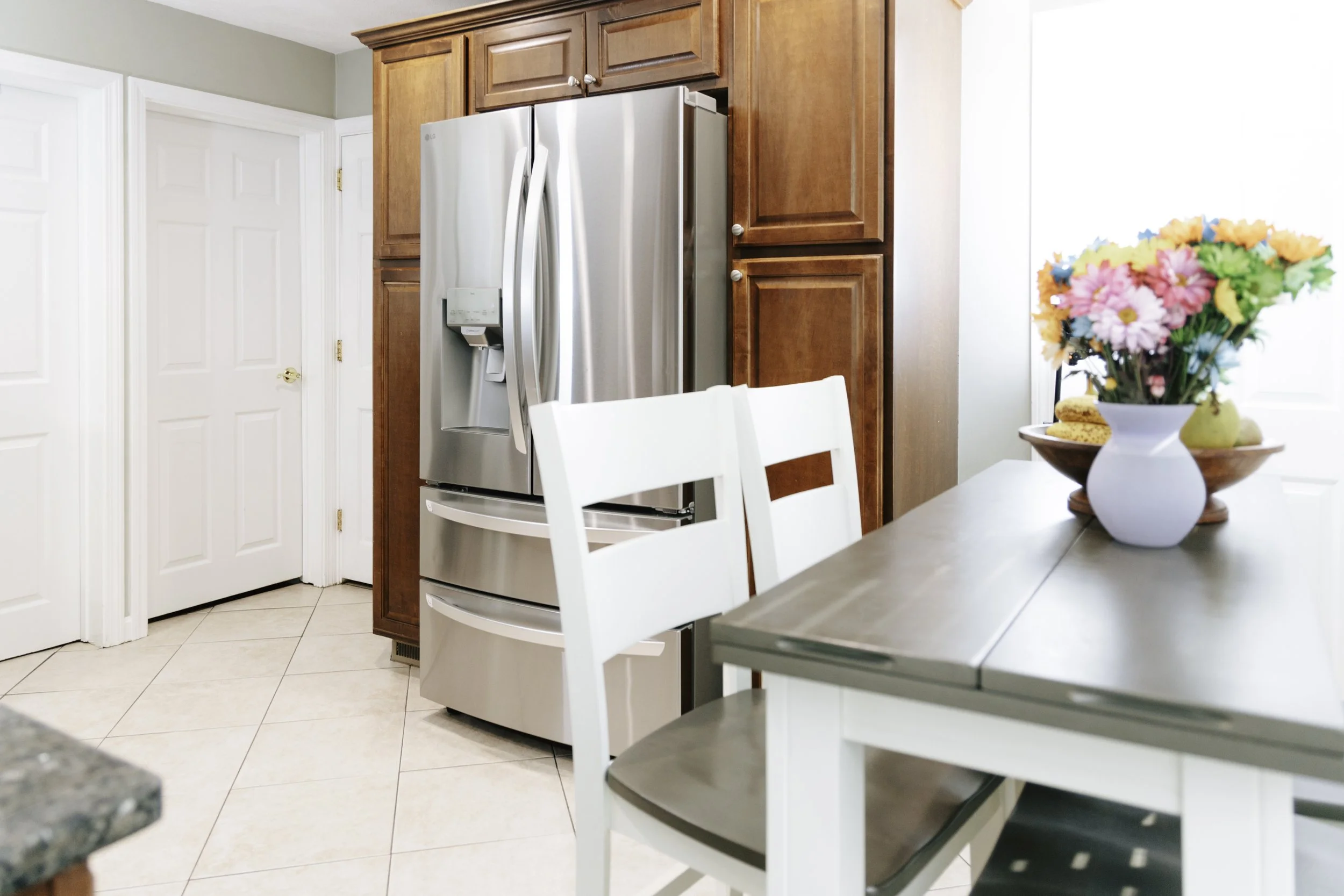 A modern kitchen with a stainless steel refrigerator, wooden cabinets, a white dining table with dark wood chairs, and a white vase with colorful flowers on the table.