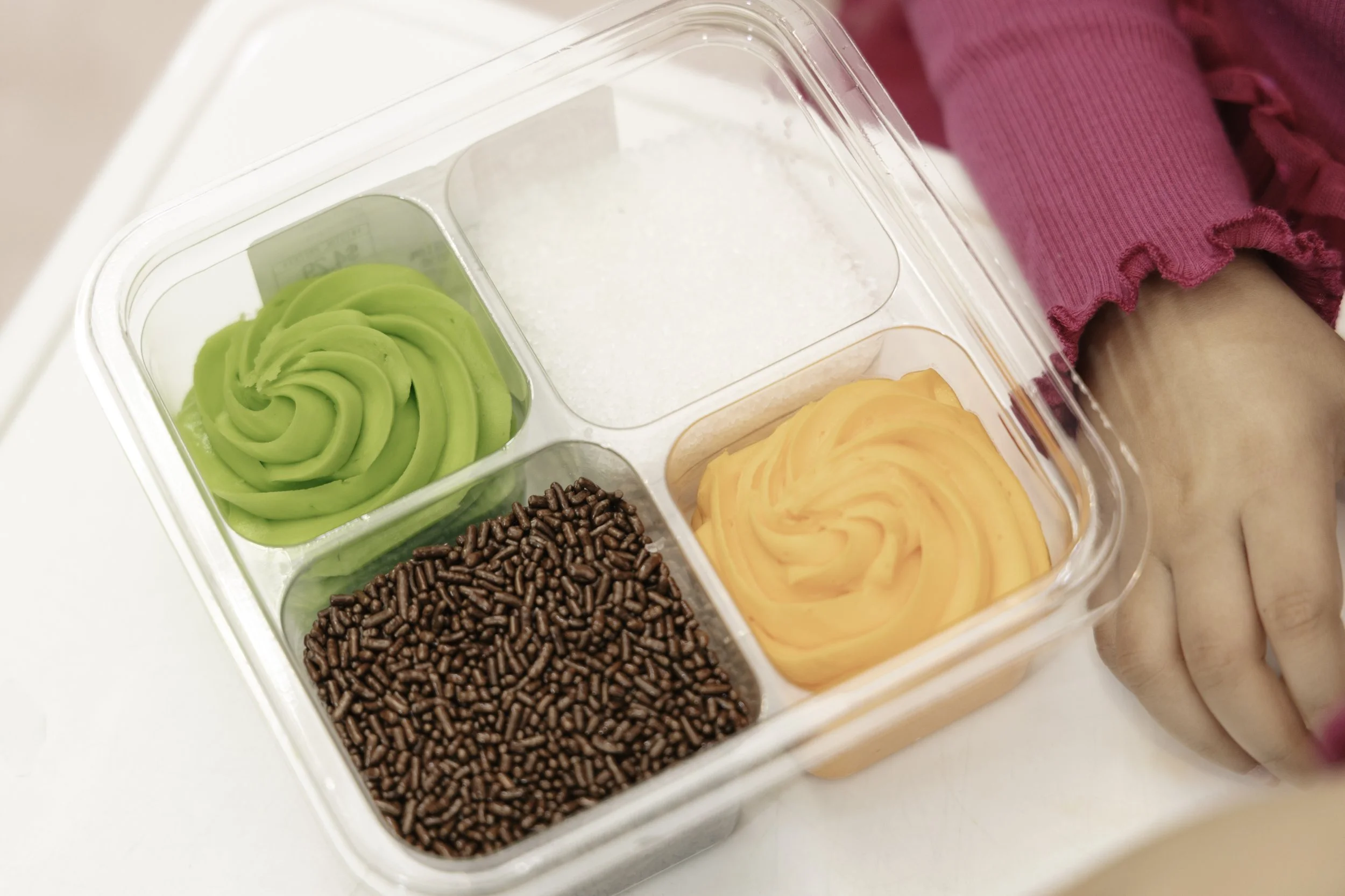 A clear plastic container with four compartments: green buttercream swirled in the top left, white sprinkles in the top right, brown chocolate sprinkles in the bottom left, and orange buttercream swirled in the bottom right. A person wearing a pink l