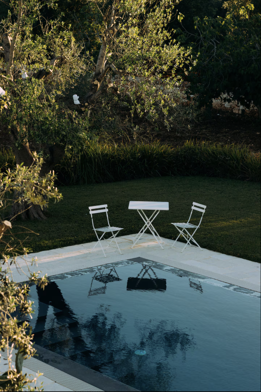 Ein Garten mit einem kleinen Pool, einem weißen Tisch und zwei weißen Stühlen vor einem bewaldeten Hintergrund.