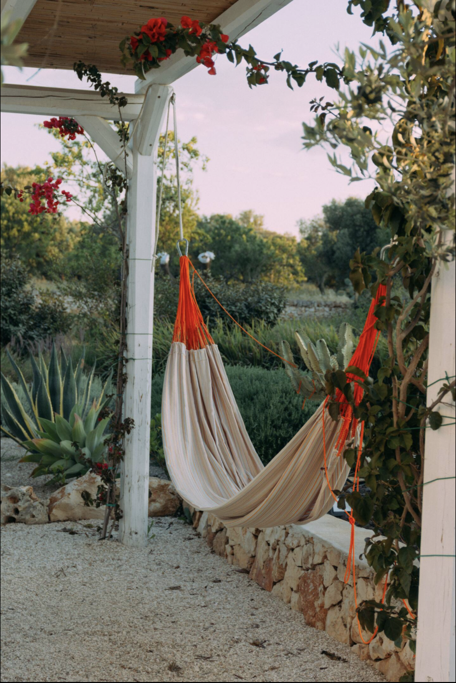 Ein Garten mit Hängesessel, Pflanzen und einem überdachten Bereich mit Holzdecke im Hintergrund, bei Sonnenuntergang.
