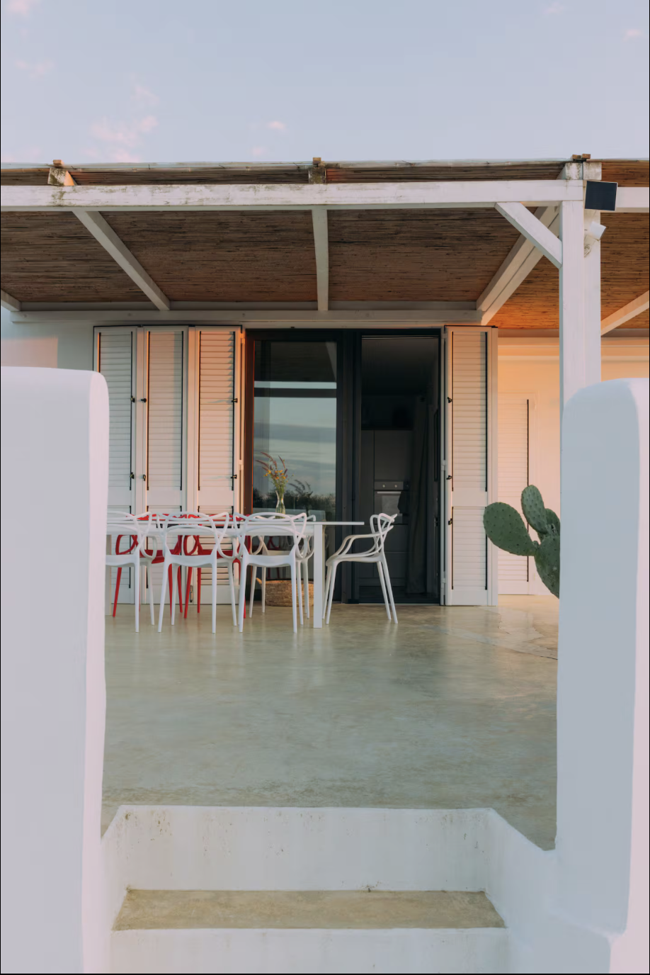 Blick auf eine Terrasse mit Esstisch und Stühlen vor einer Hausfassade mit weißen Fensterläden, eine Kaktuspflanze im Vordergrund, bei Sonnenuntergang.