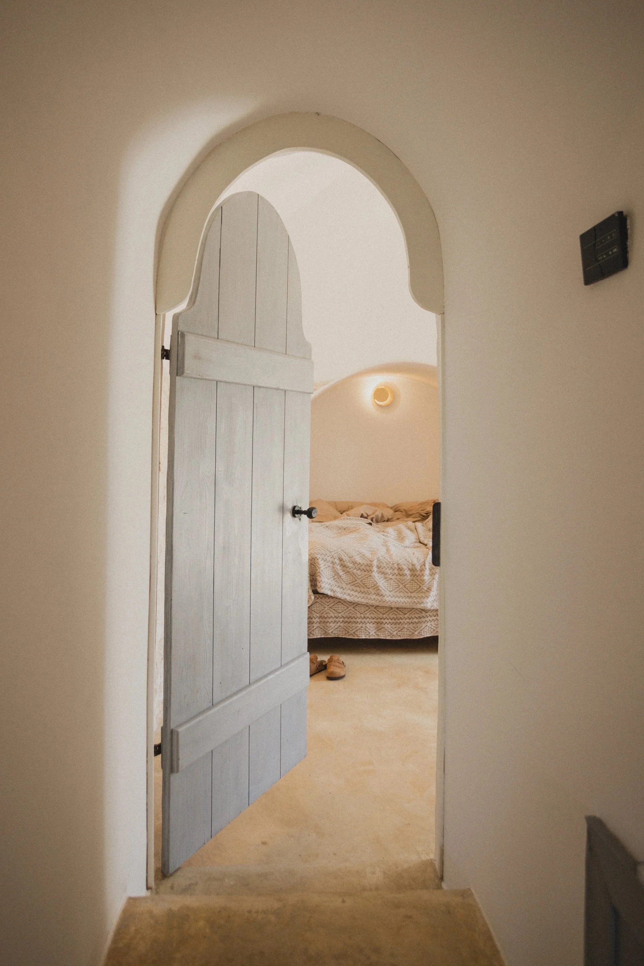 Blick durch eine offene, hölzerne Tür in ein Schlafzimmer mit einem unordentlichen Bett, eine Wandlampe und eine kleine Lampe an der Decke, im Farbton Beige gehalten.