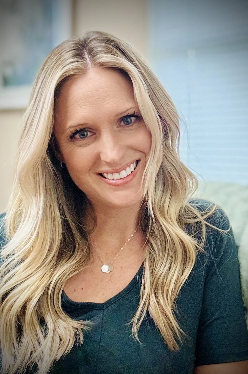 A smiling woman with long blonde wavy hair, wearing a black top and a delicate necklace, in an indoor setting with a blurred background of a window with blinds.