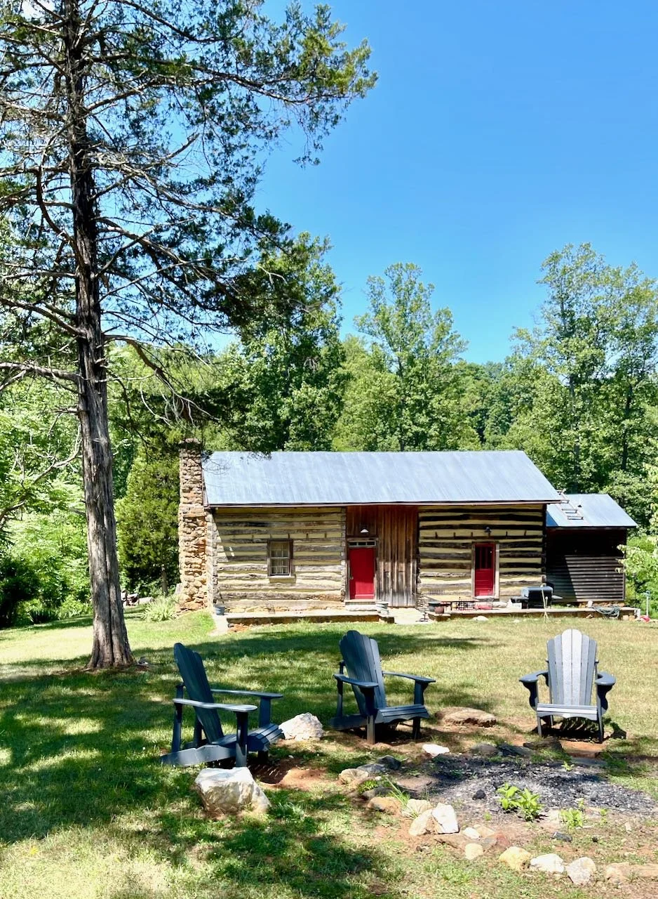 Dogtrot log-cabin with bright red doors, with a fire-pit in front.