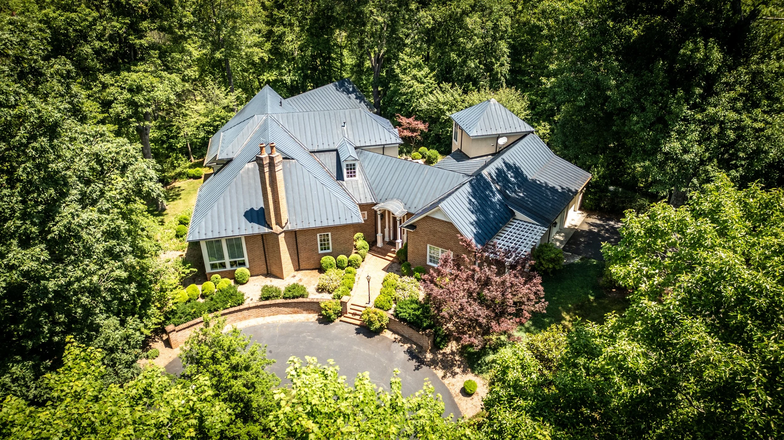 Aerial view of a large brick house with a metal roof surrounded by trees and greenery.