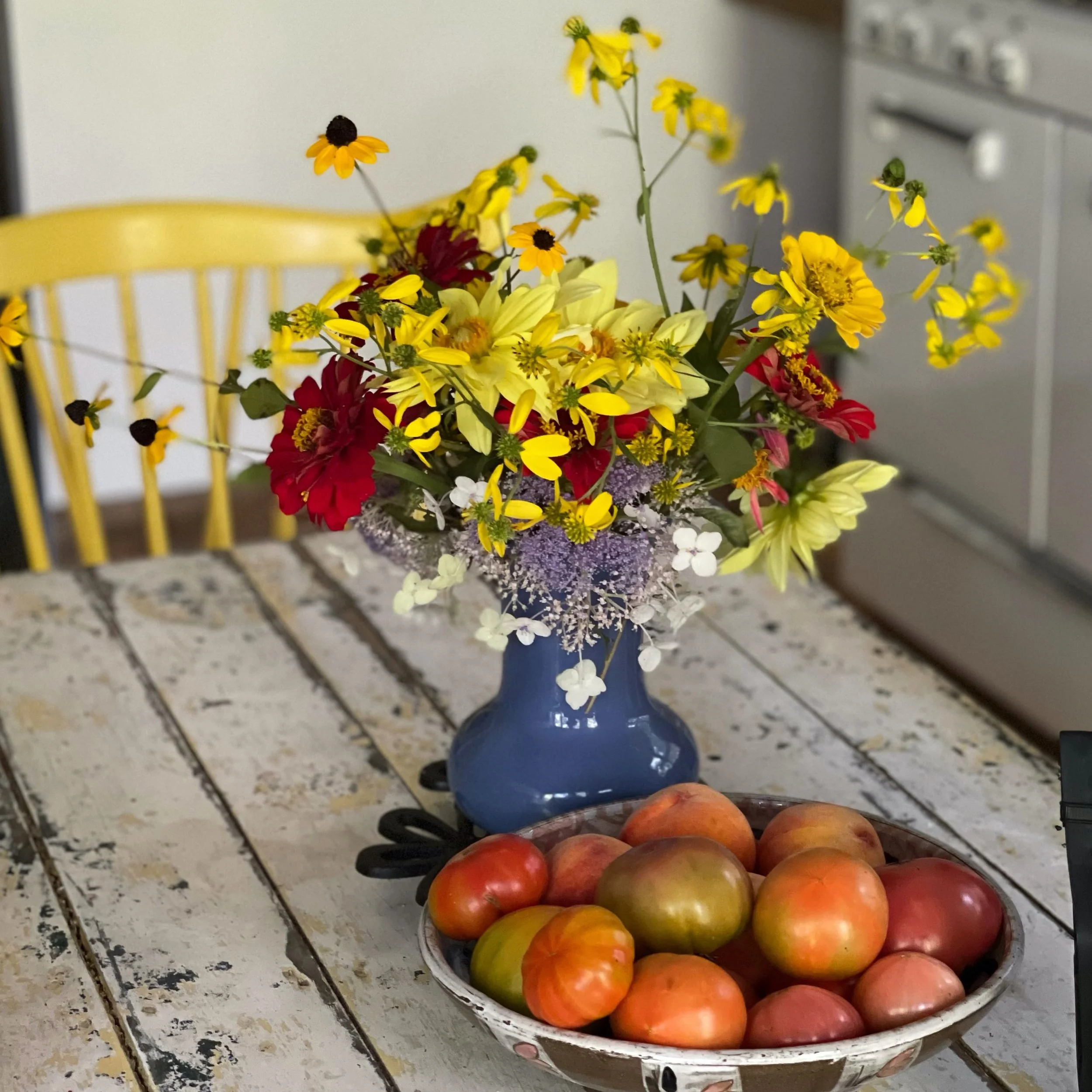 A bouquet of mixed yellow, red, and white flowers in a blue vase on a rustic white wooden table, with a bowl of ripe heirloom tomatoes in front of it and a yellow Windsor chair in the background.