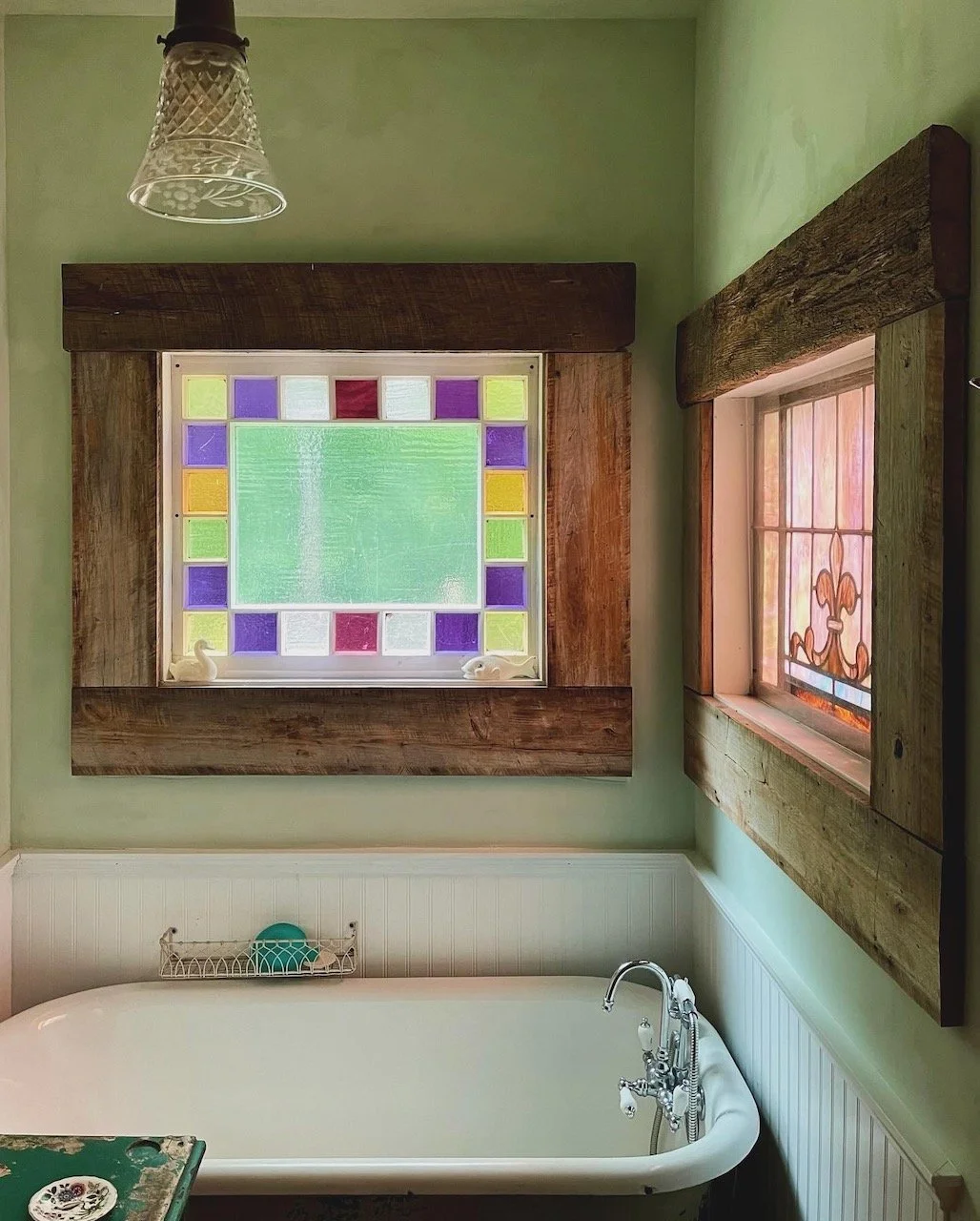A bathroom with light green walls, a white bathtub, and antique stained glass windows with wooden frames. At Spindle Hill Farm, a boutique Farm Stay in Nellysford, Virginia in Nelson County.