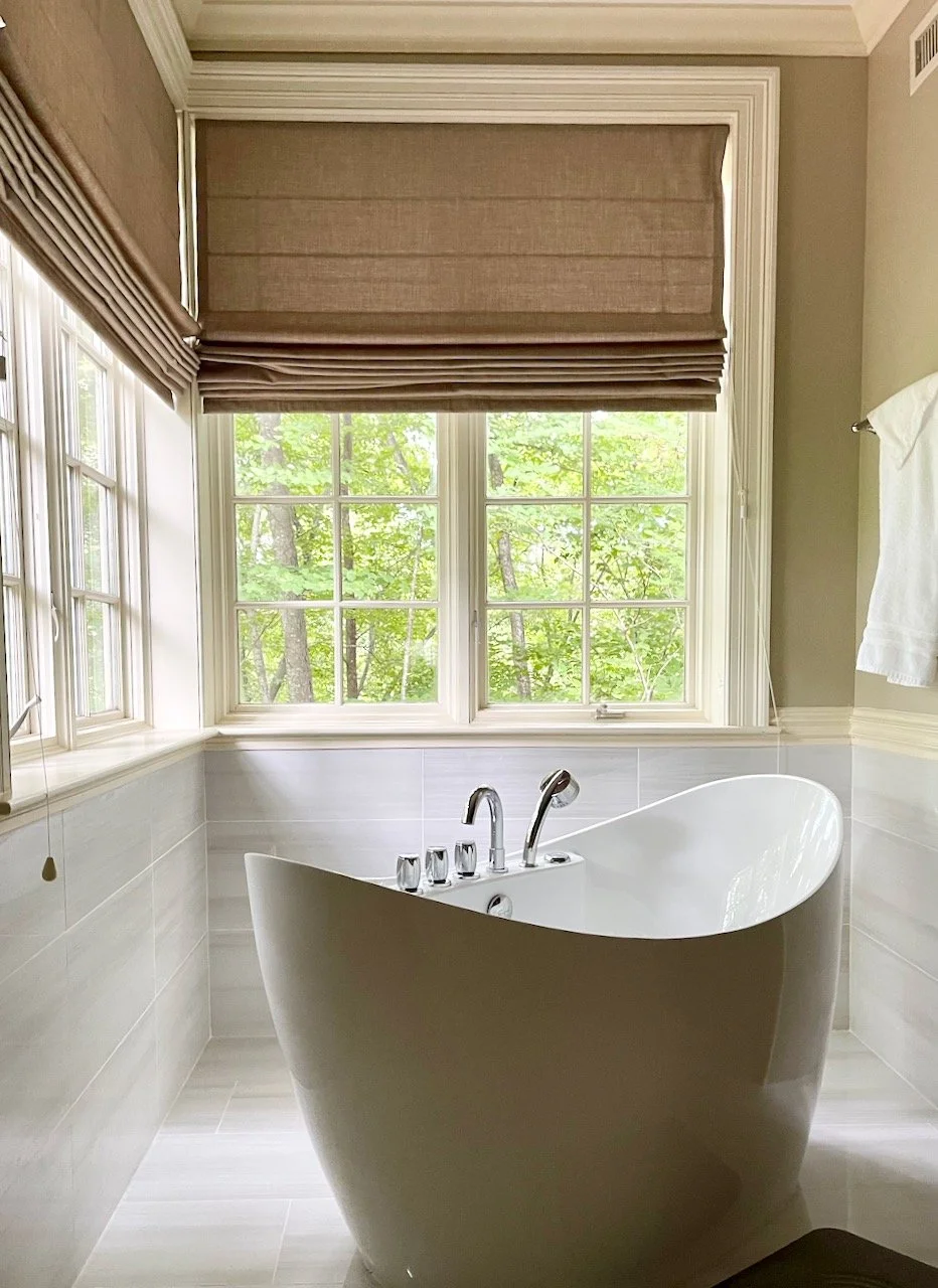A freestanding bathtub in a bathroom with large windows showing green trees outside, beige walls, and a window shade.