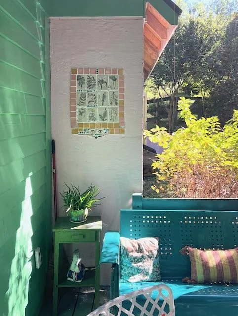 Bright outdoor patio area with green, white, and blue painted walls, a small table with a plant, a turquoise bench with pillows, and a garden view with trees and bushes.