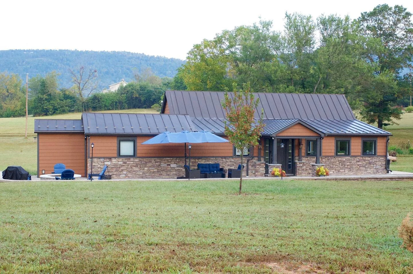 A modern single-story house with a metal roof, brown siding, stone accents, and outdoor seating area with an umbrella, set in a grassy yard with trees and distant hills.