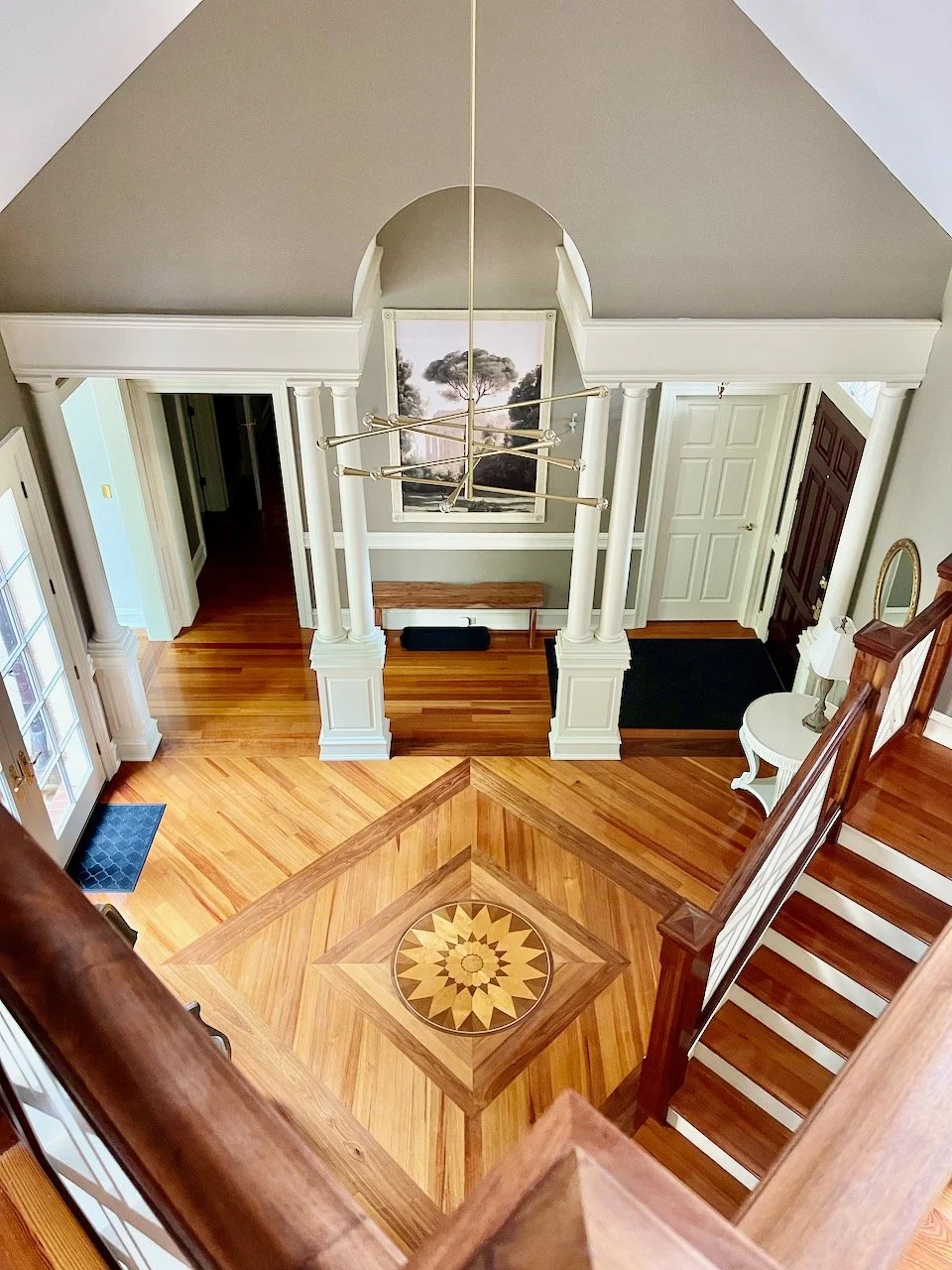 Interior view of a spacious foyer with a wooden staircase, intricate wood flooring with a geometric pattern, a chandelier, and decor including a framed picture and a small side table.