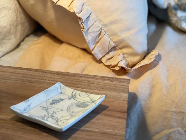 A small, white, rectangular dish with a floral design on a wooden surface next to a bed with beige pillows and a yellow blanket.