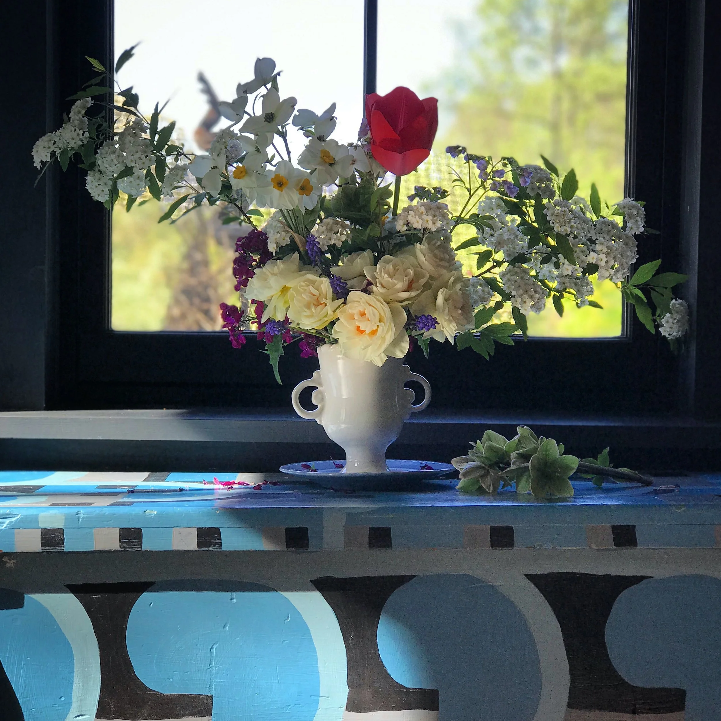 A white ceramic vase with a floral arrangement, placed on a blue and white hand-painted painted 19th Century chest near a window, with greenery outside. The art deco design and pottery at by Elise Lauterbach.