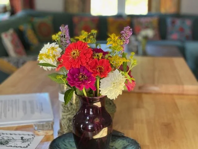 A colorful bouquet of flowers in a dark purple vase on a wooden table with papers nearby in a bright, cozy room.