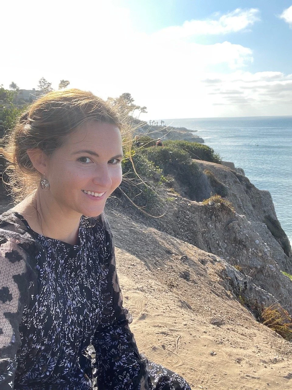 Elise Lauterbach smiling at the camera takes a selfie on a rocky cliff overlooking the Pacific ocean on a sunny day.