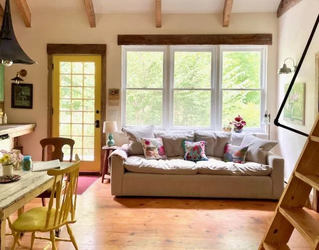 Living room with large window, beige sofa with colorful pillows, yellow door with glass panes, wooden beams on ceiling, wooden staircase, and a dining table with chairs on the left.