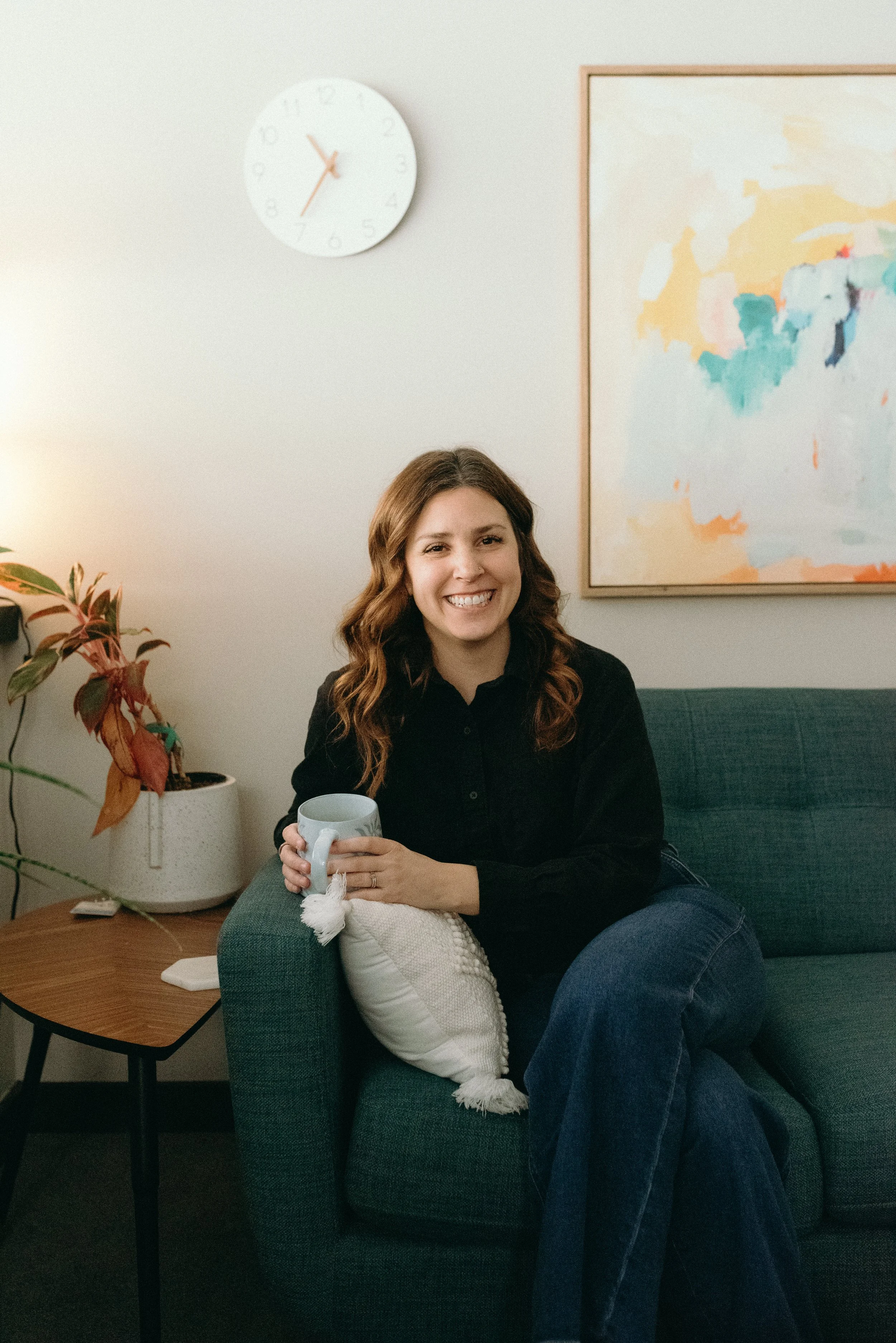 Photo of women with long brown hair smiling gleefully while sitting on a green couch and wearing a black shirt. She holds a coffee mug in her hand and a clock is behind her on the wall.