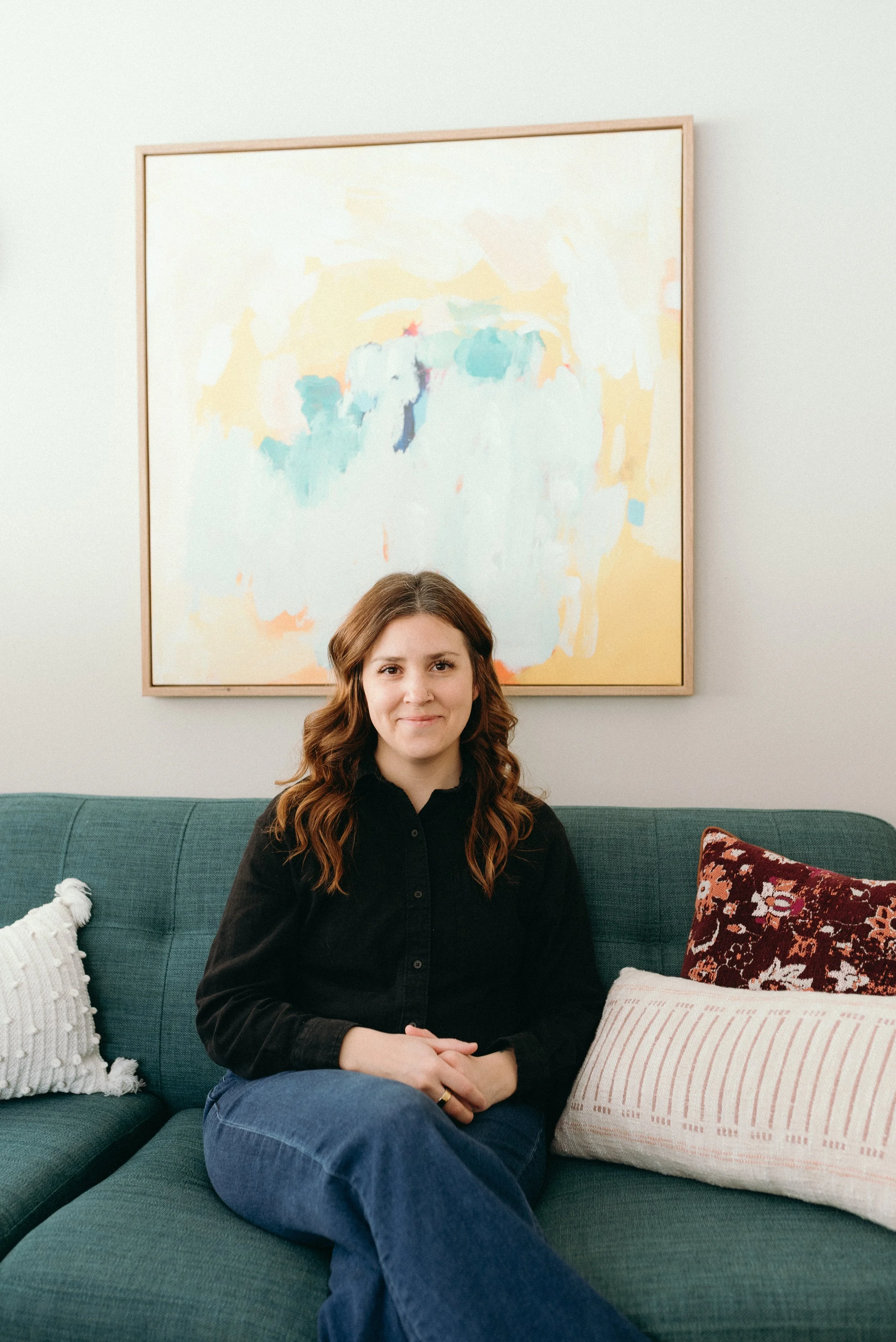 Photo of woman with long brown hair, wearing a black shirt while sitting on a green couch grinning. An abstract painting with soft and welcoming colors is on the wall behind her.